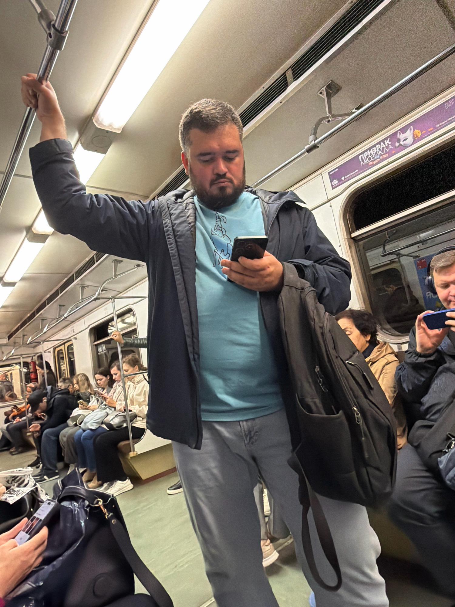 This is a medium shot taken inside a Moscow Metro train car. The primary subject is a man standing in the aisle, holding onto a vertical pole with his left hand and looking down at his smartphone. He is wearing a dark jacket over a light blue t-shirt and light grey pants. A black backpack is slung over his right shoulder. The train car is illuminated by fluorescent lights. Several other passengers are visible, some seated and others standing. In the background, there is advertising on the wall of the train car. The overall impression is that of a typical commute on public transportation.