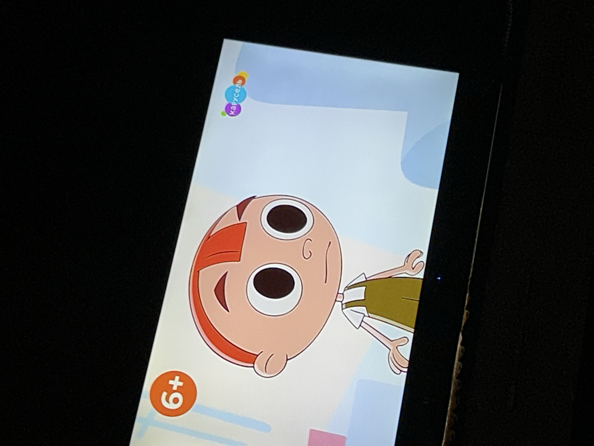 The image shows a close-up, angled shot of a television screen displaying an animated cartoon. The cartoon features a young boy with a bald head, orange hair styled in a fringe, large round eyes, and a small nose and mouth. He is wearing a white t-shirt and green overalls. In the top left corner of the screen, there is an orange circle with "6+" inside, indicating a rating. In the top right corner, there is a logo with colorful circles and the word "Карусель" (Carousel in Russian). The background of the cartoon is a light blue with abstract shapes. The television is black and appears to be placed in a dark room, with the screen reflecting the ambient light. There is no discernible information in the image that places it in Gelendzhik, Russia.