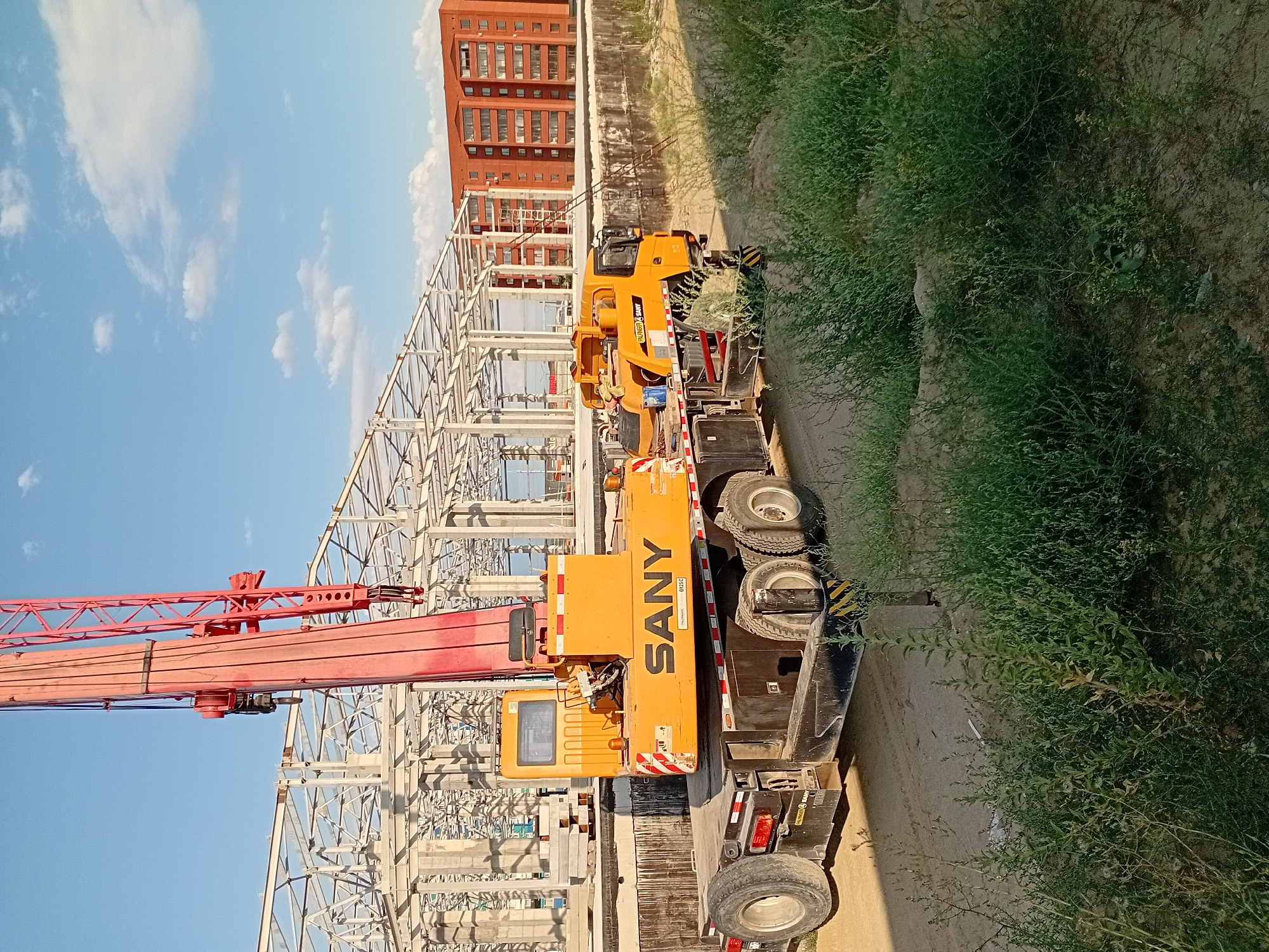 The image depicts a construction site in Semey, Kazakhstan, with a large SANY brand crane and its trailer prominently featured in the foreground. The crane, which is red and yellow, is positioned on a black trailer with multiple wheels. Behind the crane, a skeletal steel structure of a building is under construction. To the right of the construction site, a multi-story brick building is visible. The sky is blue with scattered white clouds. The ground is a mix of dirt and sand, with green vegetation growing in patches. The scene suggests ongoing construction activity in an urban or semi-urban environment. There are no people visible in the image.