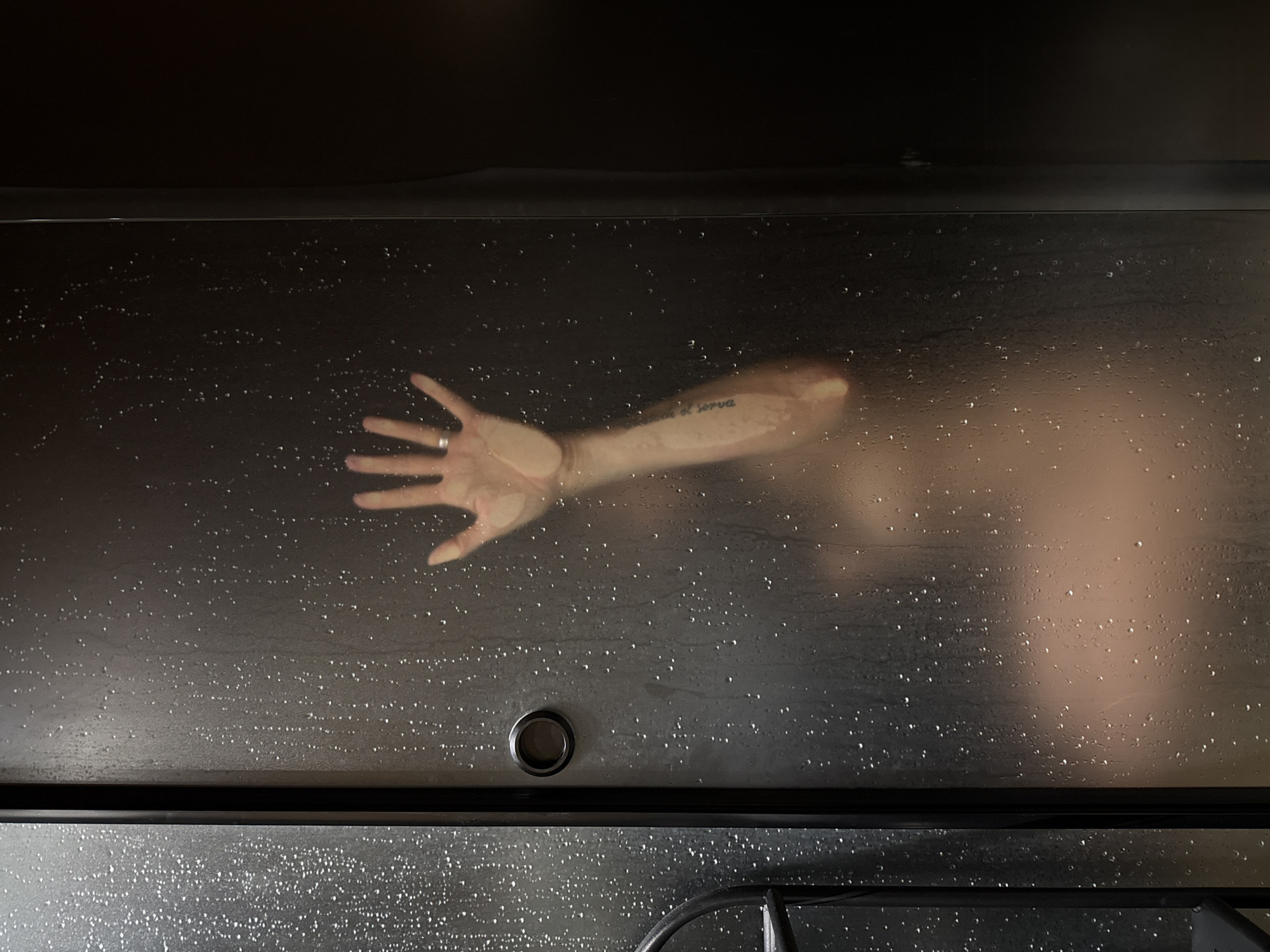 A person's hand is pressed against a frosted glass shower door, obscuring their body. The hand is raised with fingers spread, and a ring is visible on one finger. The arm has visible tattoos. Water droplets are visible on the glass, suggesting the shower is in use or has recently been used. The background is dark and indistinct. There is no information about the location in the city of Chaikovsky, Russia, provided in the image.