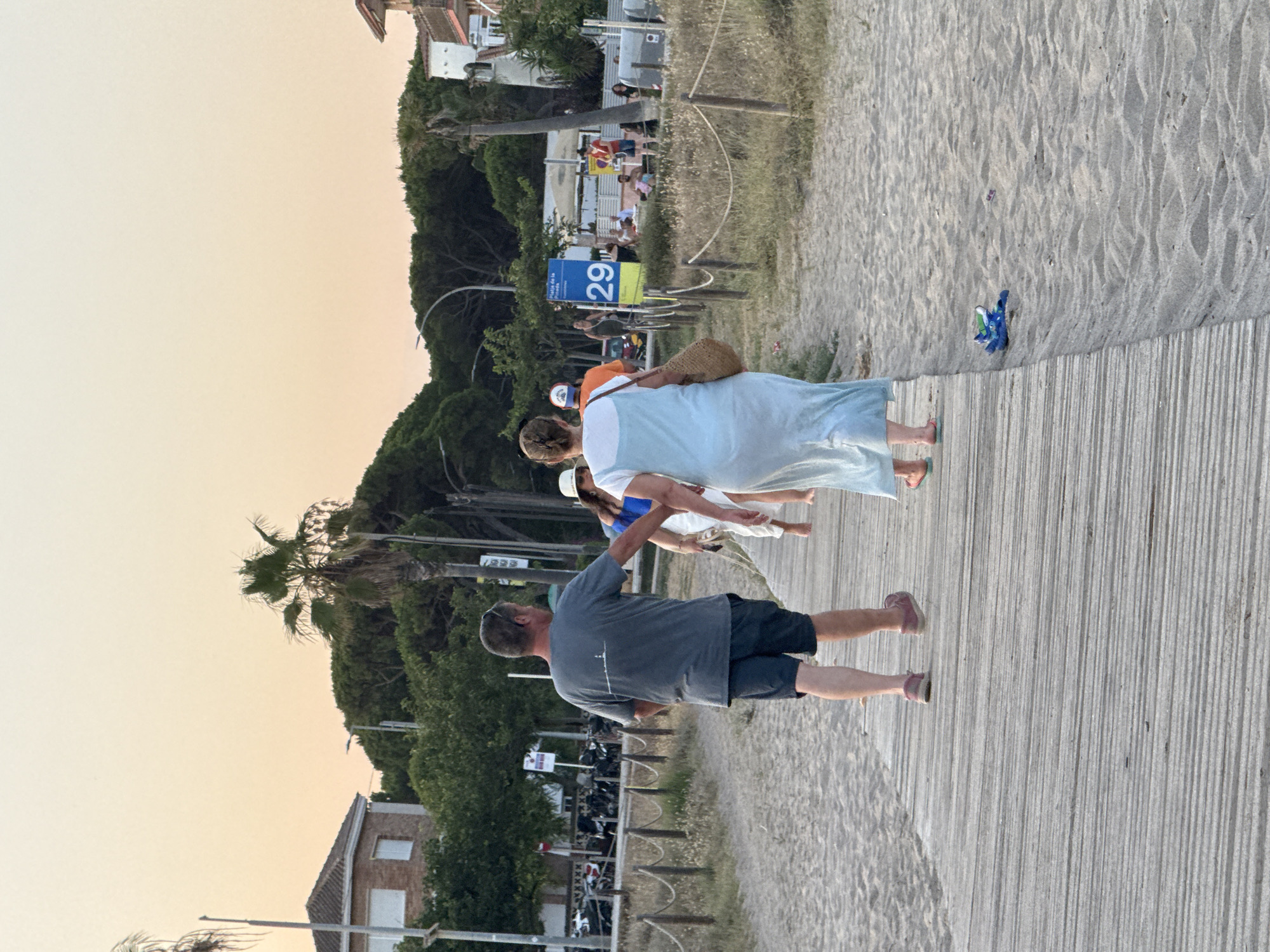 The image captures a scene on a beach boardwalk in Castelldefels, Spain, during the late afternoon or early evening. The sky is a soft, pale orange, indicative of sunset. A wooden boardwalk, composed of parallel planks, runs diagonally from the lower left to the upper right of the frame, bordered by sand on both sides.
In the foreground, a man in a grey t-shirt and dark shorts walks away from the camera on the boardwalk. Beside him and slightly ahead, a woman in a light blue, flowing dress walks in the same direction, carrying a woven straw bag. Behind them, a woman in a white hat and blue top is partially visible. Further down the boardwalk, several other people are walking and standing.
To the left of the boardwalk, there are sand dunes with sparse vegetation and a rope fence. In the background, a row of buildings, including a house with a red tile roof, are visible amongst lush green trees and palm trees. A street with parked motorcycles and a signpost with the number "29" and text in Spanish indicating "Playa de la Pineda" (Pineda Beach) are also present, confirming the location. The overall atmosphere suggests a leisurely summer evening by the sea.