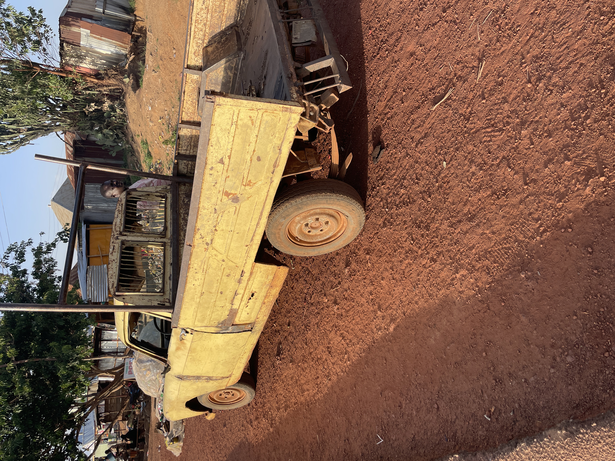 The image depicts an old, yellow pick-up truck parked on a red dirt surface. The truck appears to be in a state of disrepair with visible rust. A person is visible inside the cab of the truck. The background features trees, buildings with corrugated metal roofing, and general signs of a rural or less developed environment. The image does not contain identifiable details to place the image at Durimi, Nigeria.