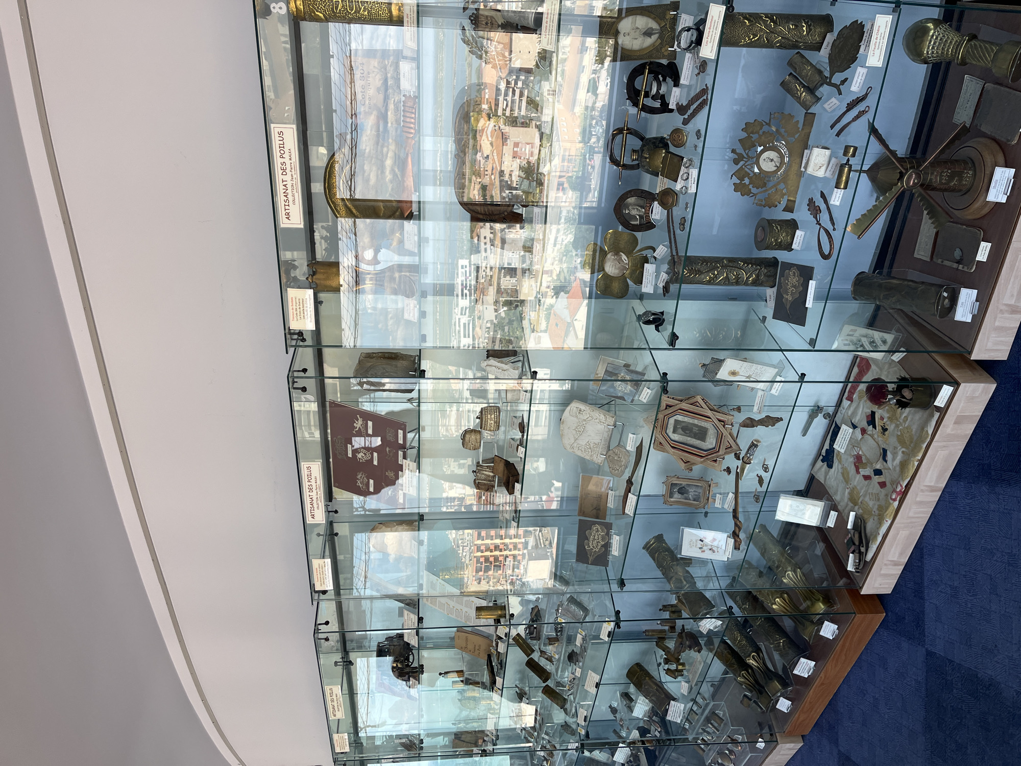 The image shows a display of historical artifacts in glass cases, likely in a museum, situated in Palavas-les-Flots, France. The display features a collection of items, possibly related to military history or everyday life from a past era, with labels indicating "ARTISANAT DES POILUS" and specific collection names like "COLLECTION Jean-Pierre MALKA." The artifacts include what appear to be brass shell casings, intricate metalwork, personal items, framed photographs, and decorative objects, many of which are ornate and handcrafted. The background visible through the glass cases suggests an elevated perspective overlooking a coastal town with buildings and the sea, consistent with the location of Palavas-les-Flots. The lighting is bright, illuminating the items within the cases, and the floor is covered with a patterned blue carpet. There are no people visible in the image.