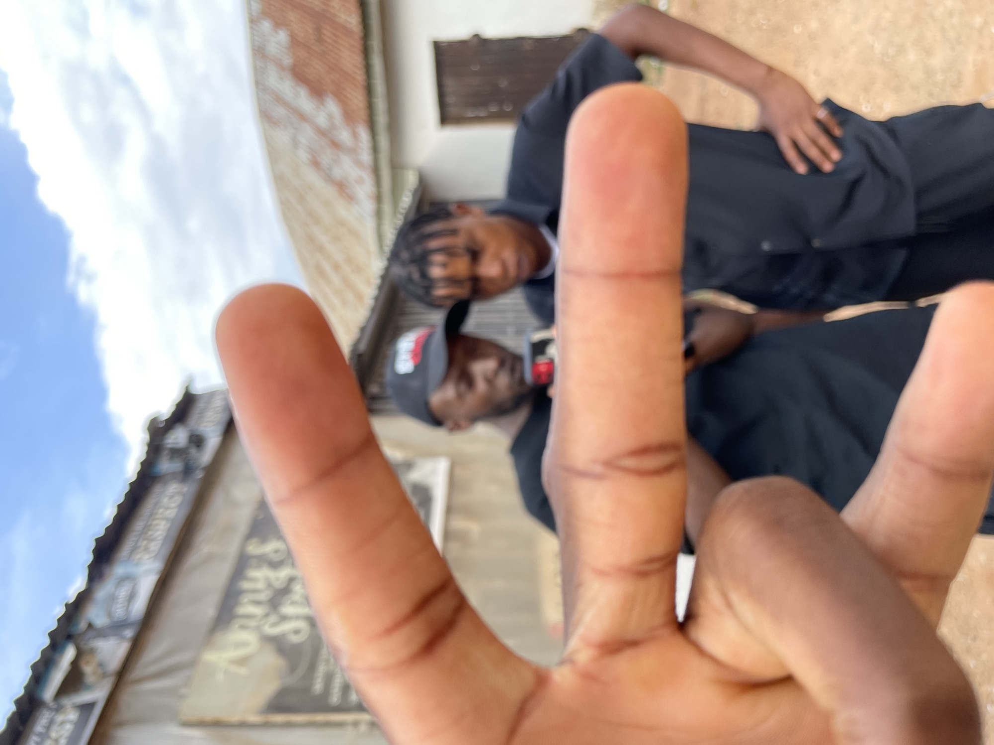 The image is a tilted, low-angle shot, likely a selfie, with a dark-skinned hand prominently positioned in the foreground, making a "V" (peace or victory) sign with the index and middle fingers extended. The hand's palm shows distinct crease lines.
Behind the hand, the scene is heavily blurred, featuring two males and an outdoor setting. The closer male, positioned to the left, wears a dark shirt and a dark baseball cap. His face is turned slightly towards the camera, but his features are indistinct due to the blur. Further back and to the right, a second male is visible, also wearing a dark shirt and sporting braided hair. His face is partially visible.
The background consists of a bright, cloudy sky on the left and various building elements on the right and bottom left. One building on the left has a visible sign. The word "RESPECT" is faintly discernible at the top of the sign, and below it, the words "Anny's Spot" or similar appear to be written. Another building section displays reddish-brown brickwork. The image conveys a casual, spontaneous moment captured during the daytime in an urban or semi-urban environment in Barakin Akawo, Nigeria.
