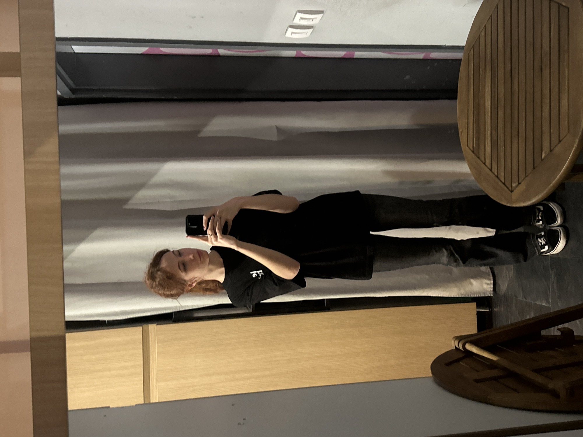 A person with auburn hair is standing in front of a mirror and taking a selfie with a smartphone. They are wearing black clothes and jeans with black and white sneakers. The reflection shows a bed with a gray headboard, a wooden table, and a wall with recessed lights. The location is likely in a residential setting in Novi Sad, Serbia.