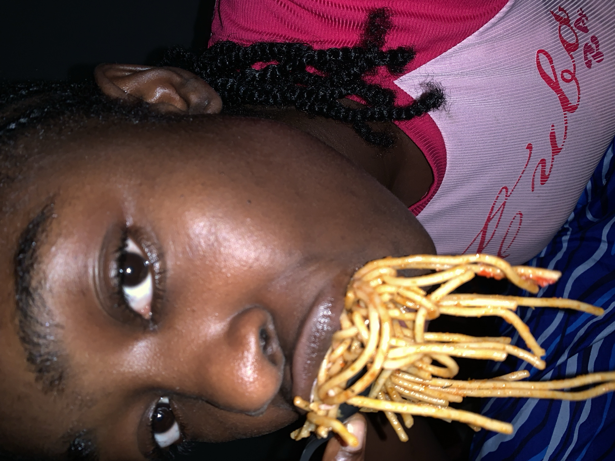 A close-up, portrait-oriented image shows a person with dark skin eating spaghetti, tilted counter-clockwise. The person is on the left side of the frame, looking towards the right. Their dark, braided or twisted hair is visible, framing their face. They wear a ribbed top that is pink or reddish on the upper section and white or light pink on the lower part, which features stylized dark red text resembling cursive, possibly "Love" or similar, though not fully legible.
A large forkful of spaghetti, coated in a reddish sauce, is being lifted towards the person's mouth, held by their partially visible right hand. The spaghetti strands are prominent in the foreground, obscuring part of the person's mouth and chin. Their eyes are open, looking directly towards the viewer, with distinct reflections of light in their irises. The ear is visible on the upper left, and the nose and lips are clearly seen.
The background on the right and bottom features a blue fabric with a lighter blue striped or patterned design, likely a blanket or another piece of clothing. The lighting suggests an indoor setting, potentially at night due to the dark background elements. The person's expression is neutral and focused on the act of eating. The scene takes place in Rafin Par, Nigeria.