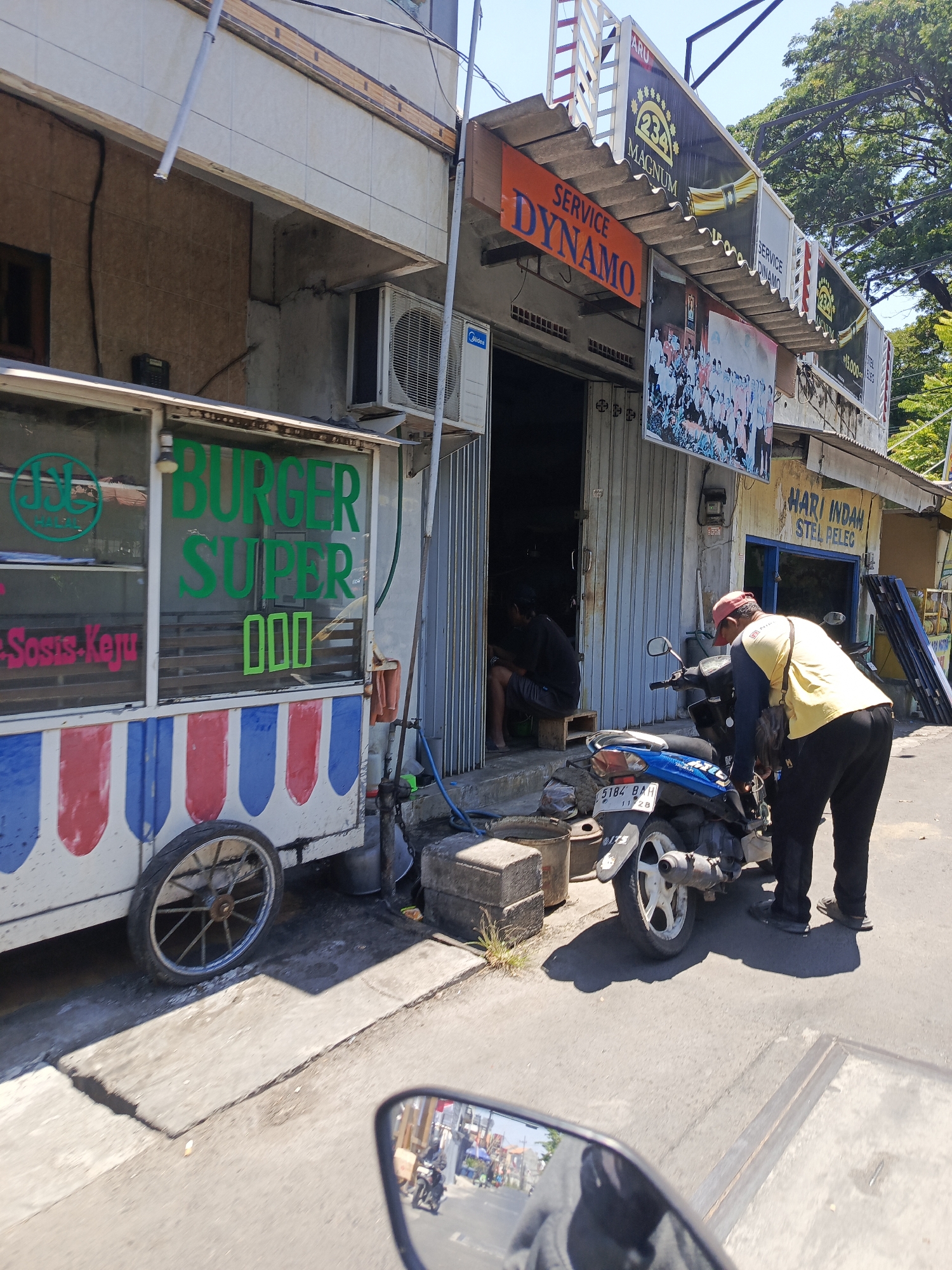 In Surabaya, Indonesia, a sunny street scene unfolds with a burger cart and a service shop. The burger cart, decorated with red and white stripes, displays green text reading "BURGER SUPER" and a halal logo. Adjacent to it, a shop with a metal roll-up door displays signs for "SERVICE DYNAMO" and "HARI INDAH STEL PELEC". A man in a yellow shirt and red cap is attending to a blue motorcycle parked in front of the shop. Another person is seated inside the shop's entrance. The ground is paved with concrete, showing cracks and wear. The reflection in a motorcycle's rearview mirror shows a street with other motorcycles and buildings.