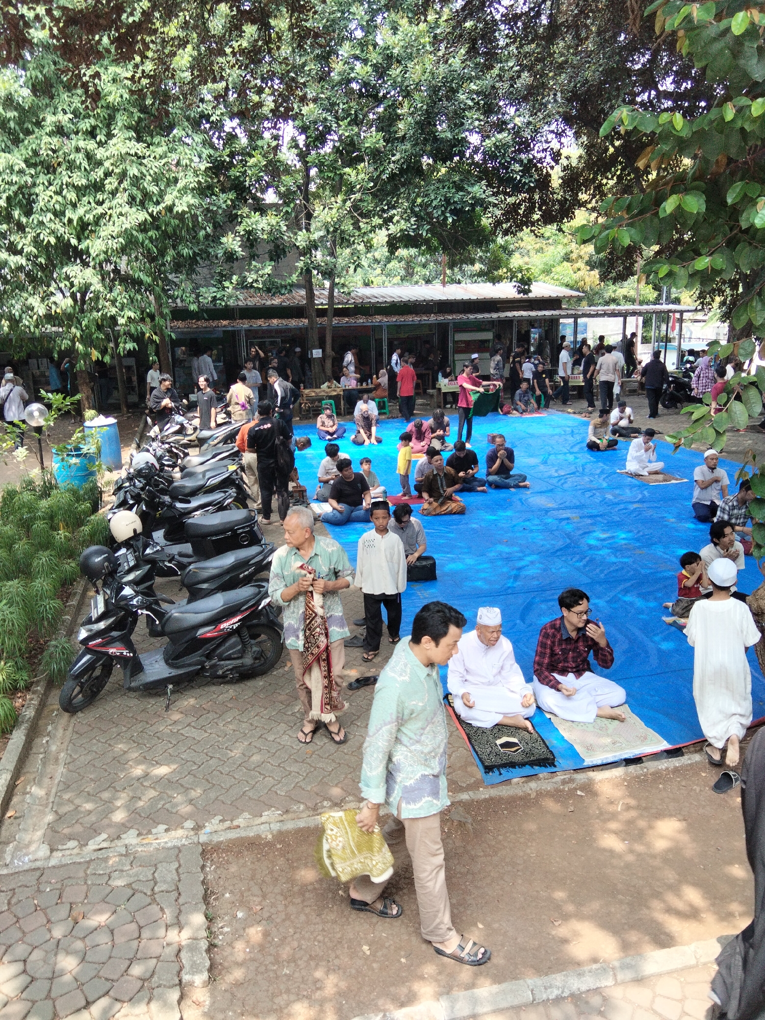 In Depok, Indonesia, under the dappled shade of large trees, a gathering of people is taking place outdoors. The scene is set on a paved area adjacent to a building with an open-air facade, and a large blue tarp spread out on the ground. Several motorbikes are parked neatly in a row on the paved area.
On the blue tarp, a group of men are seated, some in prayer positions on prayer mats, others conversing. They are dressed in traditional Muslim attire, including peci caps and sarongs. One man, in a light green patterned shirt and brown pants, stands and carries a patterned cloth bag. Another man, older and wearing a light green shirt and sarong, also stands, holding a similar cloth.
The environment is verdant, with lush green foliage from the trees overhead and a hedge to the left. The building in the background appears to be a simple structure, possibly a canteen or communal space. The overall atmosphere suggests a relaxed, community gathering, possibly associated with a religious event or social occasion.