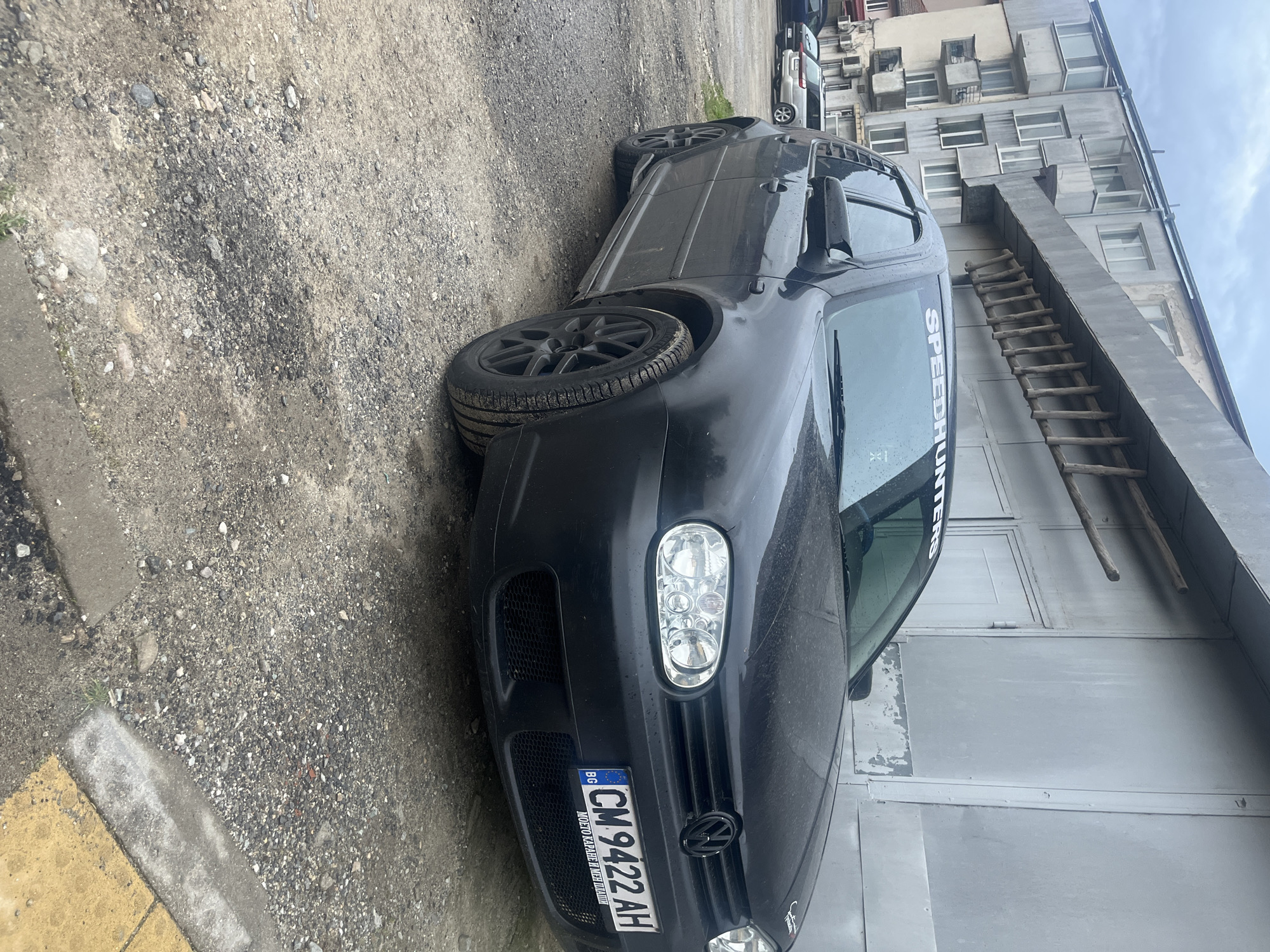 The image depicts a black Volkswagen Golf parked on a gravel and dirt surface in Zlatograd, Bulgaria. The vehicle has a "SPEEDHUNTERS" decal on the windshield and a Bulgarian license plate that reads "CM 9422 AH." The car is parked beside a light gray building with a wooden ladder leaning against the building. In the background, a multi-story residential building with numerous windows and balconies is visible. The lighting suggests an overcast day. No people are visible in the image.