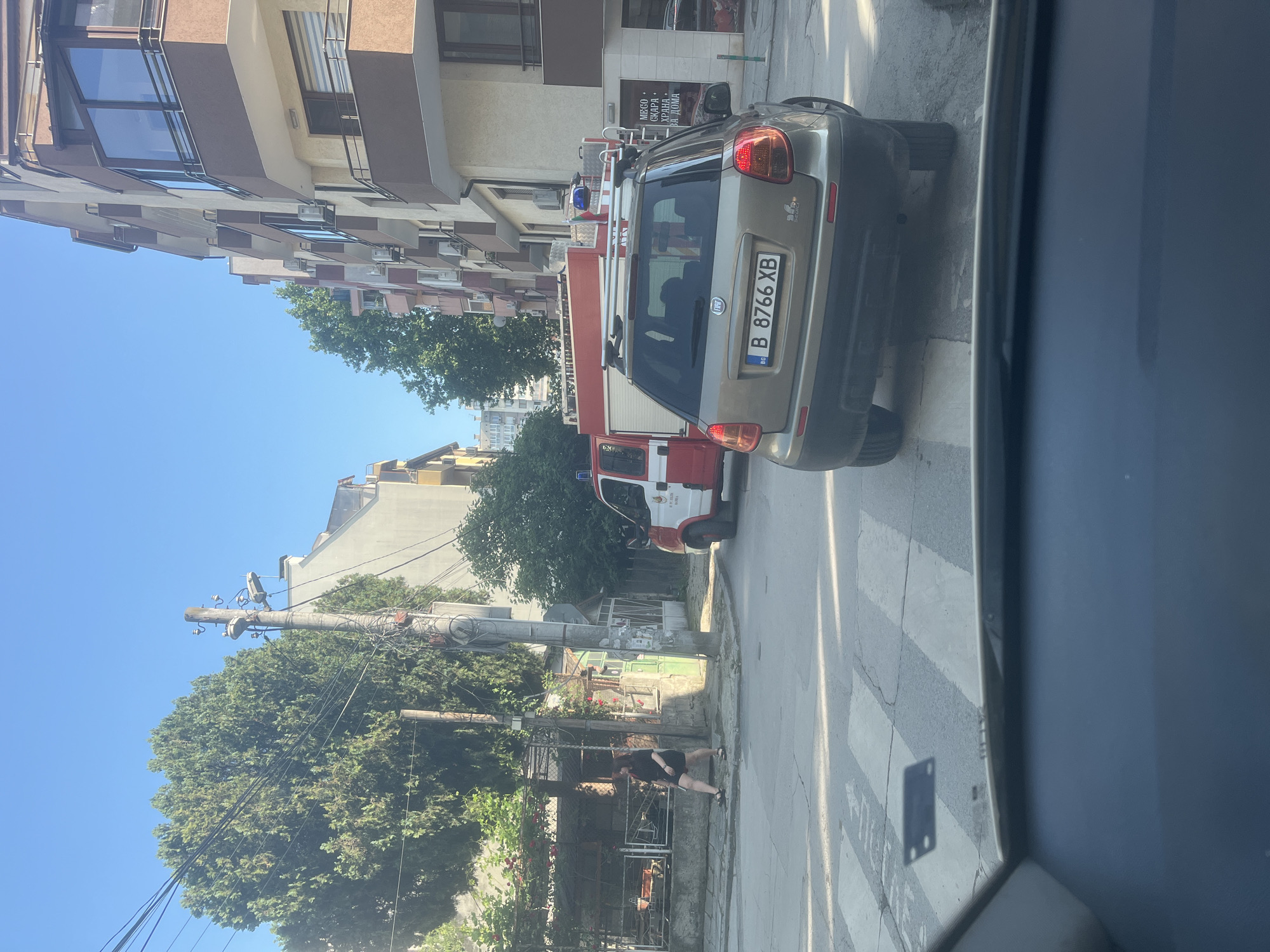 The image captures an urban street scene on a bright, sunny day, likely in Varna, Bulgaria, as provided by the context. The photograph is taken from inside a vehicle, looking out.
In the foreground, a gray Fiat Sedici car is parked on the street. It features roof rails and a Bulgarian license plate "B 8766 XB" with the "BG" national identifier. Directly behind the Fiat, partially visible, is a red and white fire truck with a blue emergency light mounted on its roof. Both vehicles appear stationary.
To the left of the parked vehicles, a person, possibly a woman, dressed in a dark t-shirt and dark shorts, stands on the sidewalk. They are positioned near a low fence or wall, next to some green foliage.
The background consists of several multi-story buildings, characteristic of residential or commercial architecture. Some buildings have balconies, windows with frames, and visible air conditioning units. On a building behind the fire truck, a sign with Cyrillic text is visible, reading: "МЕСО • СКАРА • КРЪЧМА ЗА ДОМА" (MEAT • GRILLED • TAVERN FOR HOME).
Utility poles heavily laden with electrical wires run along the street, surrounded by lush green trees and bushes, indicating a well-vegetated urban environment. The sky is clear and blue, suggesting pleasant weather conditions, likely mid-day given the bright illumination and distinct shadows. The road surface shows clear white markings of a pedestrian crosswalk.