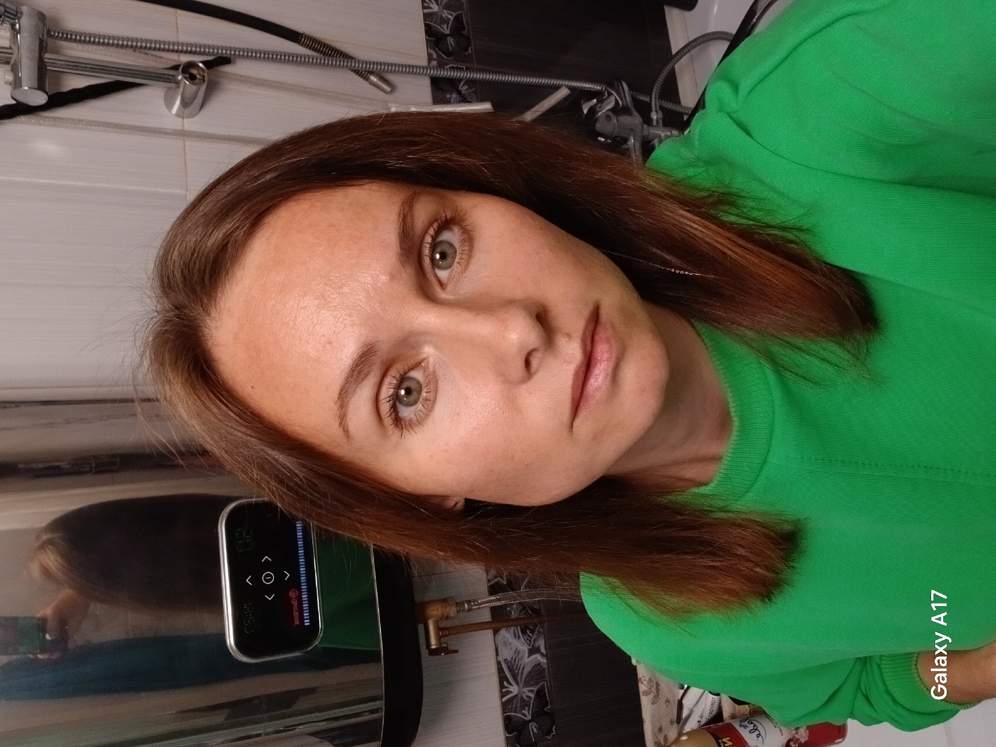 The image is a selfie of a light-skinned woman with brown hair and green eyes wearing a bright green sweater. She is indoors, in what appears to be a bathroom. Behind her is a water heater with a digital display and controls, along with a shower head and plumbing fixtures. The background walls have a combination of white tile and dark tile with a floral pattern. The picture was taken with a Samsung Galaxy A17. There is no verifiable information that would link this location specifically to Yekaterinburg, Russia.