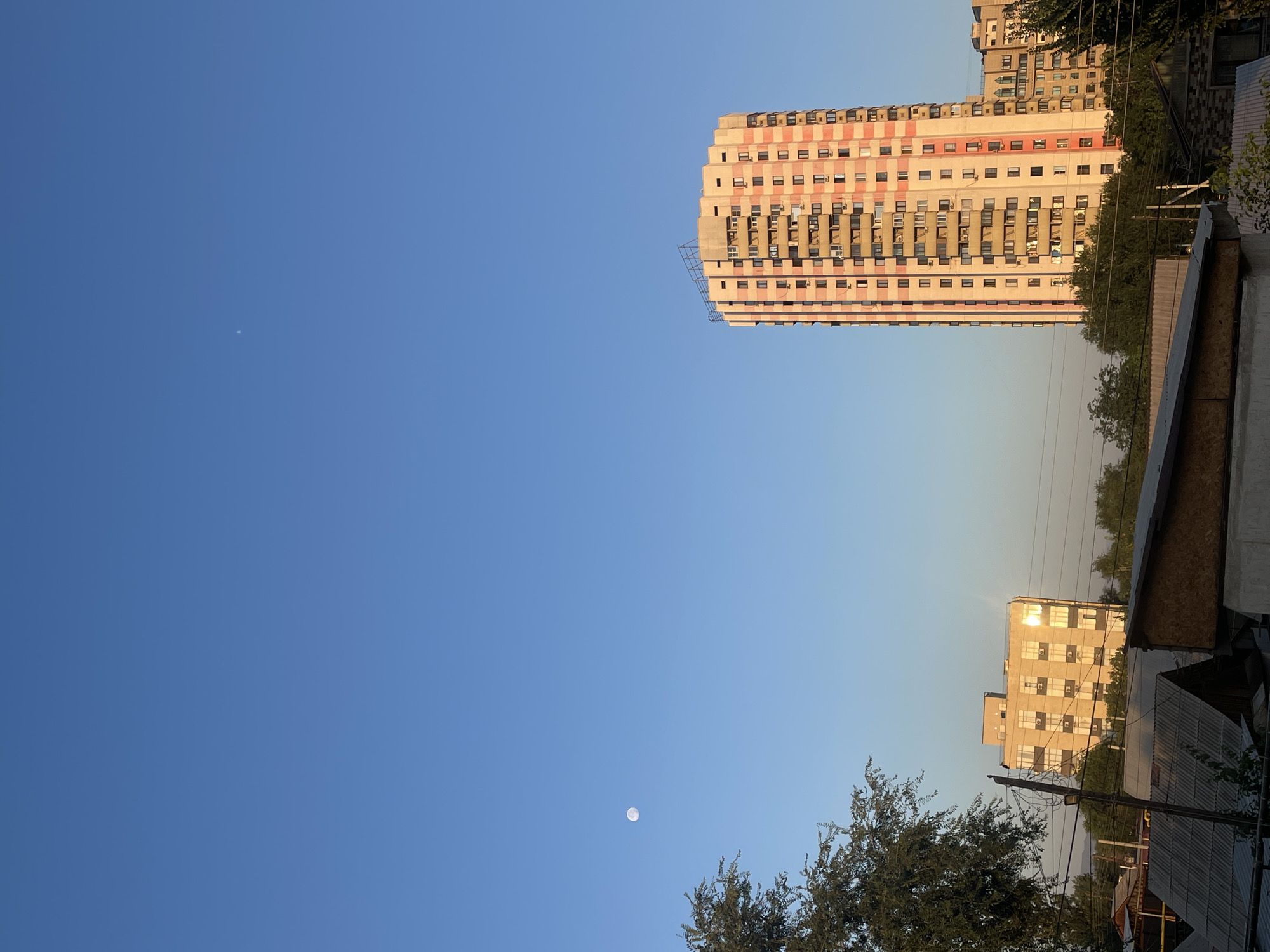 The image captures a clear, bright blue sky over a city scene in Almaty, Kazakhstan. On the right side, a tall, multi-story apartment building with a distinctive pink and cream-colored facade stands prominently. Its many windows are visible, some reflecting the sunlight. To the left of this building, a shorter, lighter-colored building with a similar grid of windows is seen, also reflecting the sun. In the far distance, more buildings and trees are visible. In the lower foreground, the roofs of several low-rise structures, possibly garages or sheds, are present. A large tree with dense foliage partially obscures the left side of the image. High in the sky, a faint full moon is visible, and a small, distant light source, possibly a planet or a distant airplane, can be observed to the upper right of the moon. The overall impression is a bright, daytime cityscape bathed in sunlight.
