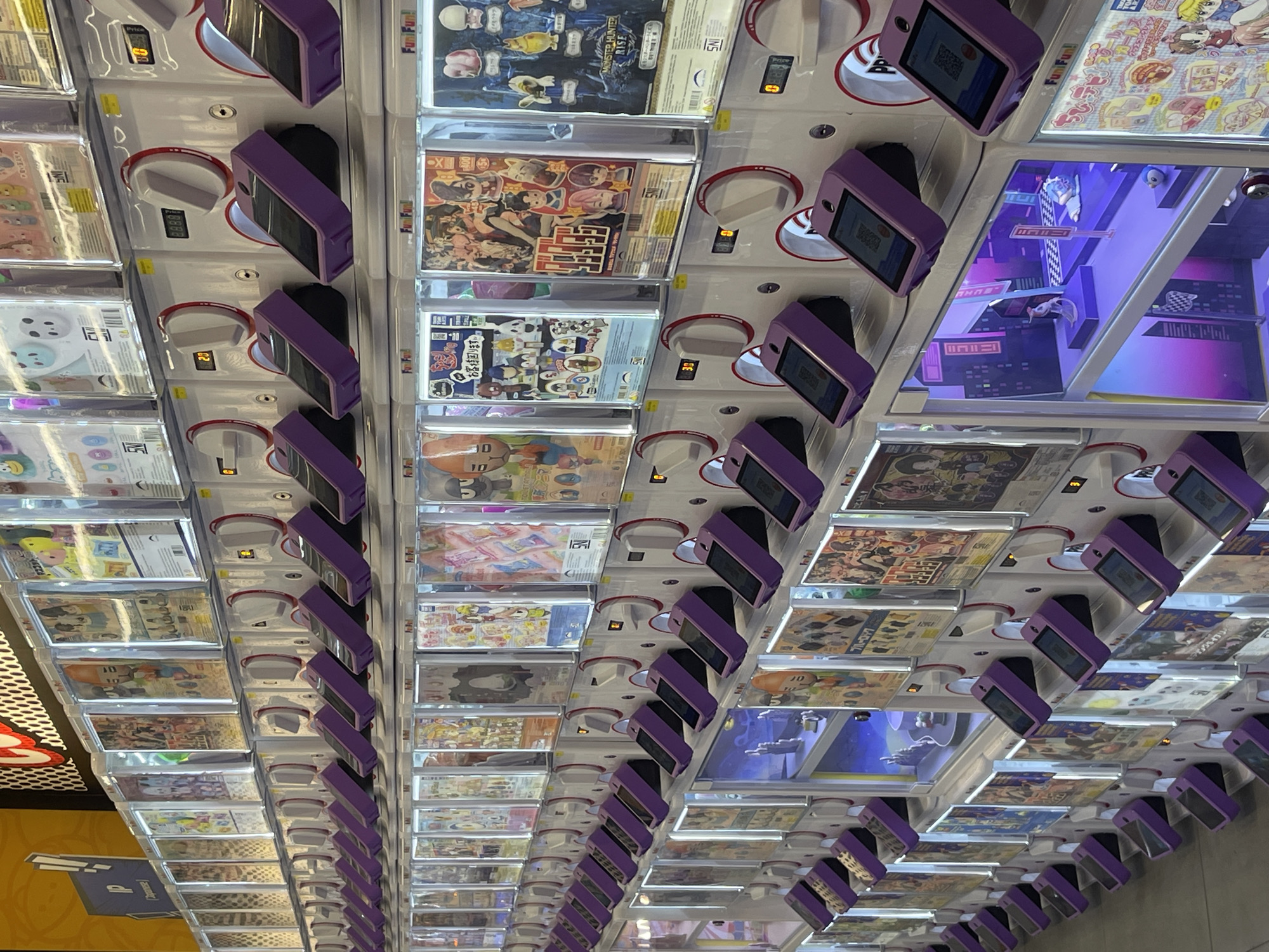 The image shows a row of gachapon machines in a store. The machines are filled with various toys and trinkets, each displayed behind a clear plastic window. The machines are white with purple levers and digital displays showing numbers. Some of the displays show the numbers "30" and "38". The gachapon machines are arranged in multiple rows, with the top rows displaying colorful images of anime characters and toys. The bottom rows have individual compartments, some of which are lit up with purple and pink lights, revealing miniature scenes with figurines. The overall environment appears to be an indoor retail space, possibly a shopping mall or a dedicated gachapon store. The image is taken from a slightly elevated angle, looking down at the machines. The city of Denpasar, Indonesia, is mentioned as the location, suggesting this is a commercial establishment within that city.