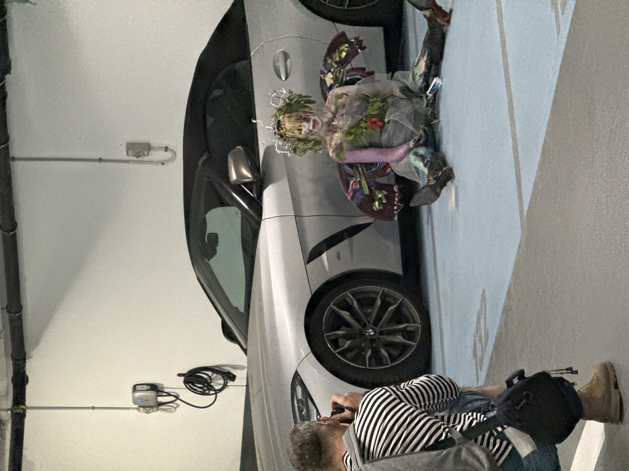 A man is photographing a person in costume leaning against a silver BMW. The location appears to be an underground parking garage. The person in costume is wearing a floral headpiece and colorful clothing with what appears to be vines or leaves as part of the costume. The man taking the photo is wearing a striped shirt, jeans, and tan boots, and he has a black camera bag near him. There is a charging station for electric vehicles mounted on the wall behind the car. It is not possible to determine the specific location in Tel-Aviv based on this image alone.
