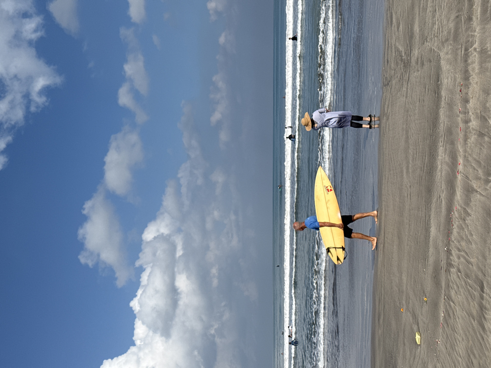 A bright, sunny day at Kuta Beach, Indonesia, features a calm ocean with gentle waves and a wide, sandy shore. In the foreground, a man in a blue shirt and black shorts walks along the wet sand, carrying a yellow surfboard under his left arm. To his right and slightly further back, a person wearing a straw hat and a long, light blue dress over black leggings stands facing the ocean, their back to the camera. Several other individuals are visible in the water, some on surfboards and others swimming. The sky is a vibrant blue, dotted with fluffy white clouds. The sand in the foreground shows subtle ripples from the receding tide, with a few small, colorful objects scattered on its surface.