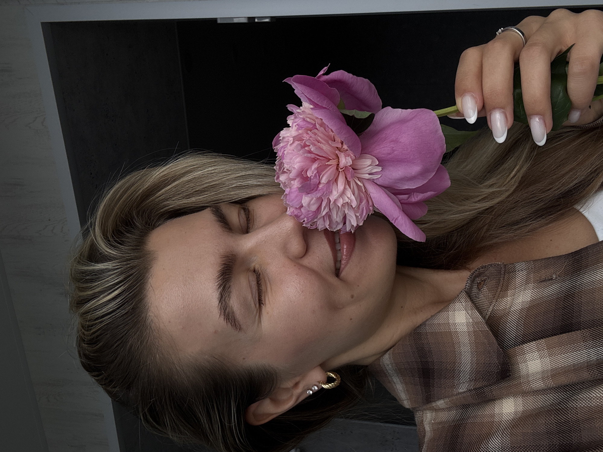 A blonde-haired individual, cropped from the mid-torso up, is depicted with closed eyes and a subtle smile, smelling a pink peony flower. The individual wears a brown and beige plaid collared shirt over a white garment. In the left ear, a small stud earring with two clear stones and a larger gold-toned twisted hoop earring are visible. The left hand, adorned with a silver-toned ring on the ring finger, holds the green stem of the peony. Fingernails feature a French manicure style with natural bases and white tips. The background is a shallow depth of field, revealing a light gray textured wall on the left and top, transitioning to a dark gray or black surface with white trim on the right, indicative of an indoor setting. The location is an unknown indoor space in the city of Unknown, Unknown.