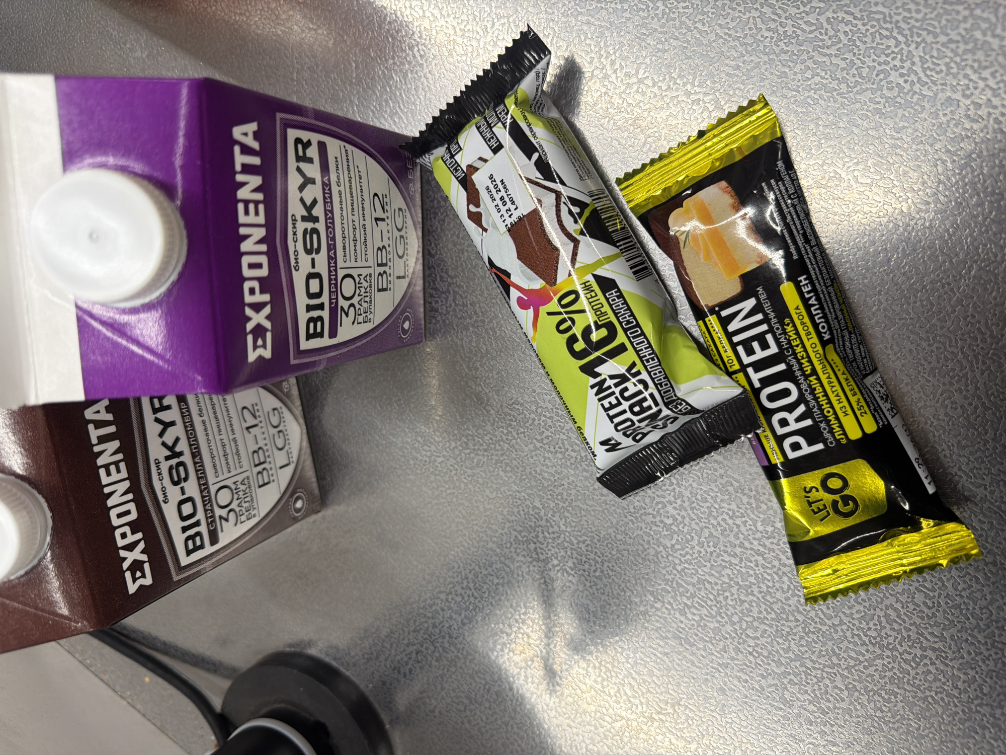 The image shows a flat, metallic surface with several food items placed on it. There are two rectangular cartons labeled "EXPONENTA BIO-SKYR," one with a brown color scheme and the other in purple, both indicating "30 GRAMM BELKA" (30 grams of protein) in Russian. Two protein bars are present. One is a lime green and white bar labeled "PROTEIN 16%," and the other is a yellow and black bar labeled "PROTEIN LET'S GO." The protein bars and drinks appear to be some kind of snack for fitness or nutrition purposes. All the packaging on all items are in Russian language and they appear to be laid on a countertop in the city of Krasnoyarsk, Russia.