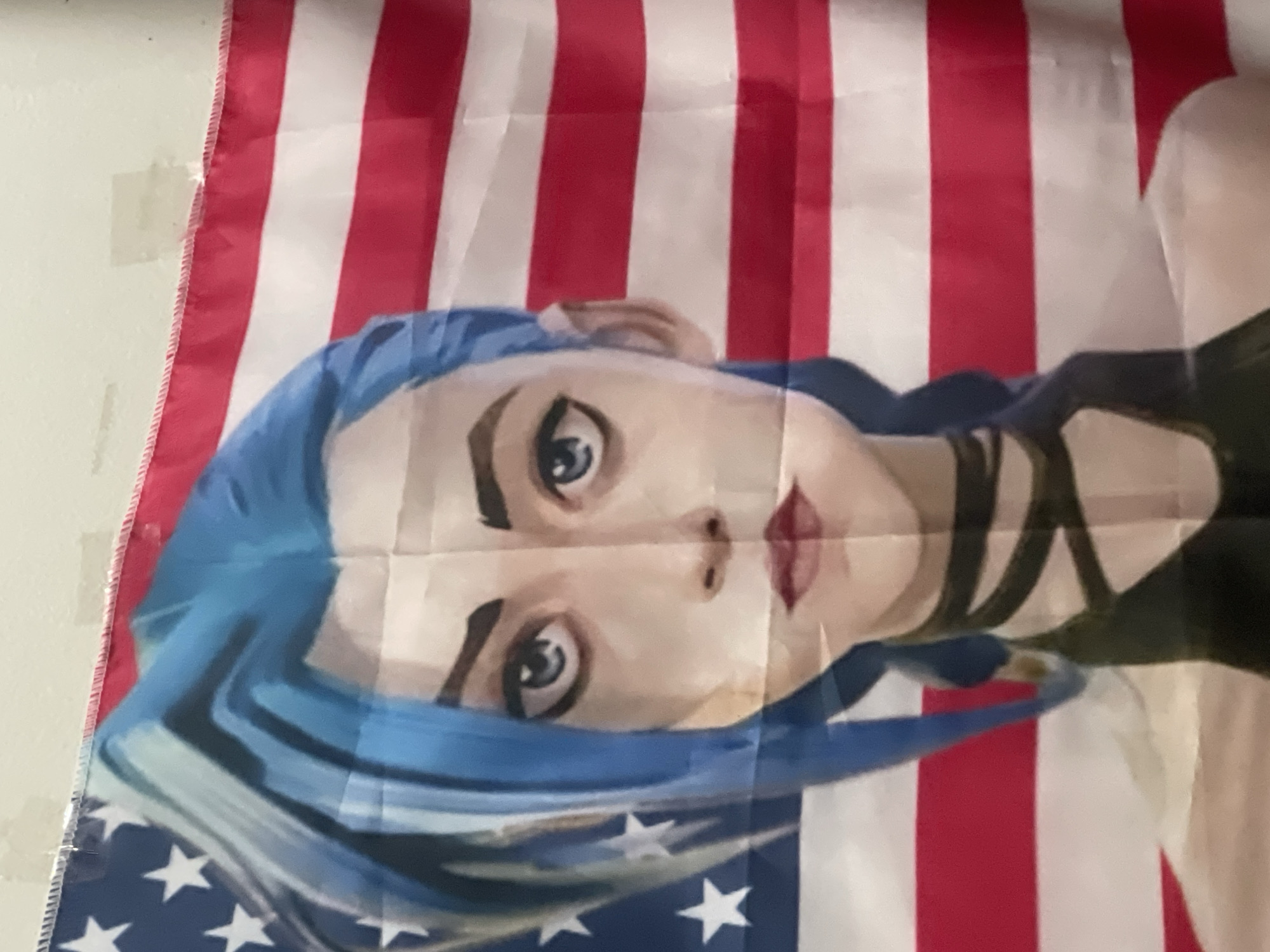 A close-up shot of a custom flag featuring a representation of the character Jinx from the animated series "Arcane" overlaid on an American flag. The flag displays red and white stripes and a blue field with white stars. The image is taken indoors, with a plain white wall visible in the background. There is no evidence to indicate a location in Denpasar, Indonesia.