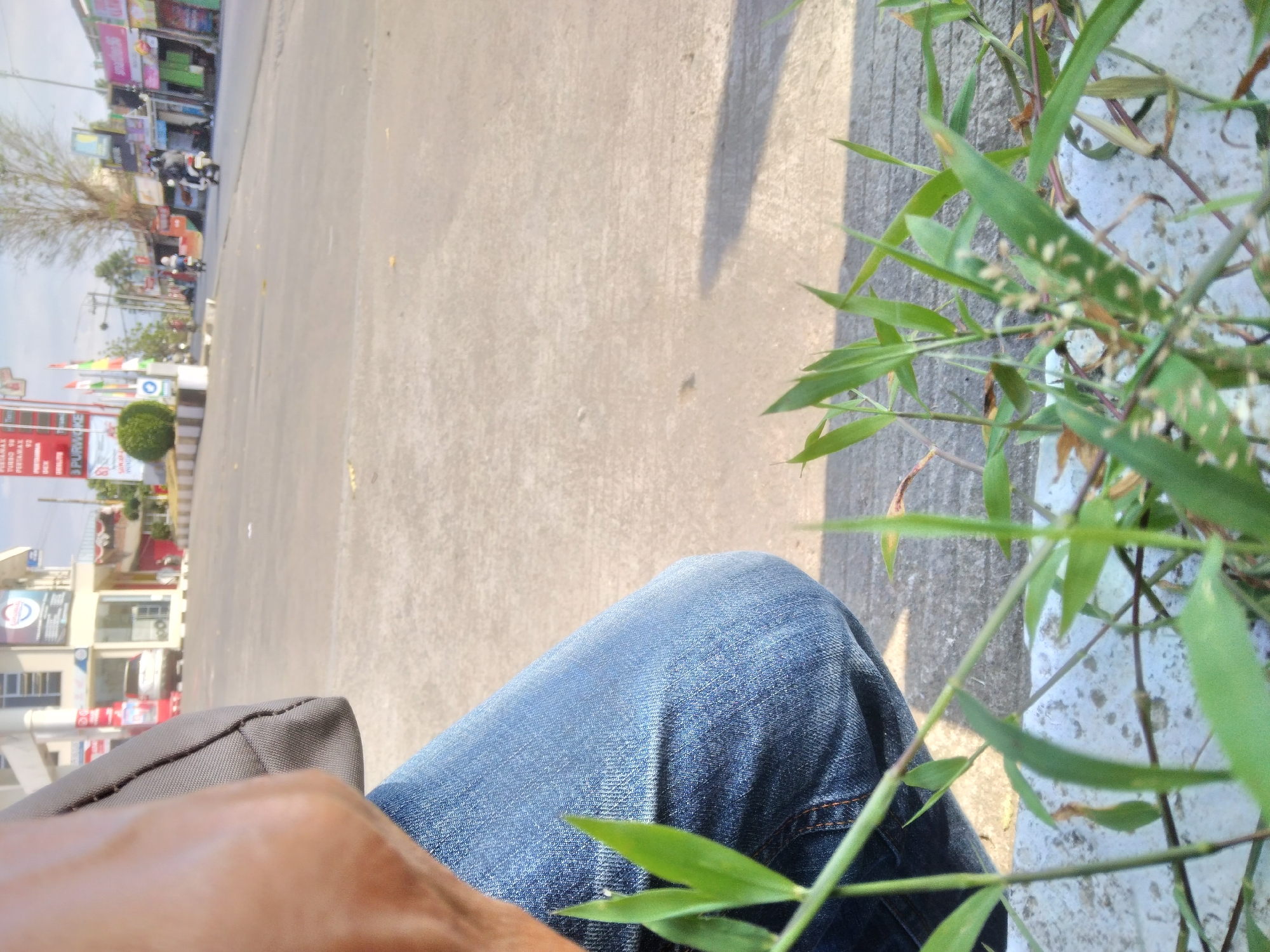 A first-person perspective shot captures a person squatting near a street in Purwokerto Kidul, Indonesia. The focus is on the person's blue jeans and a gray bag slung over their shoulder, with the foreground filled with lush green grass. In the background, a street scene unfolds with shops, buildings, and passing motorcycles under a bright sky. The overall impression is of a casual moment on a sunny day in a bustling Indonesian town.
