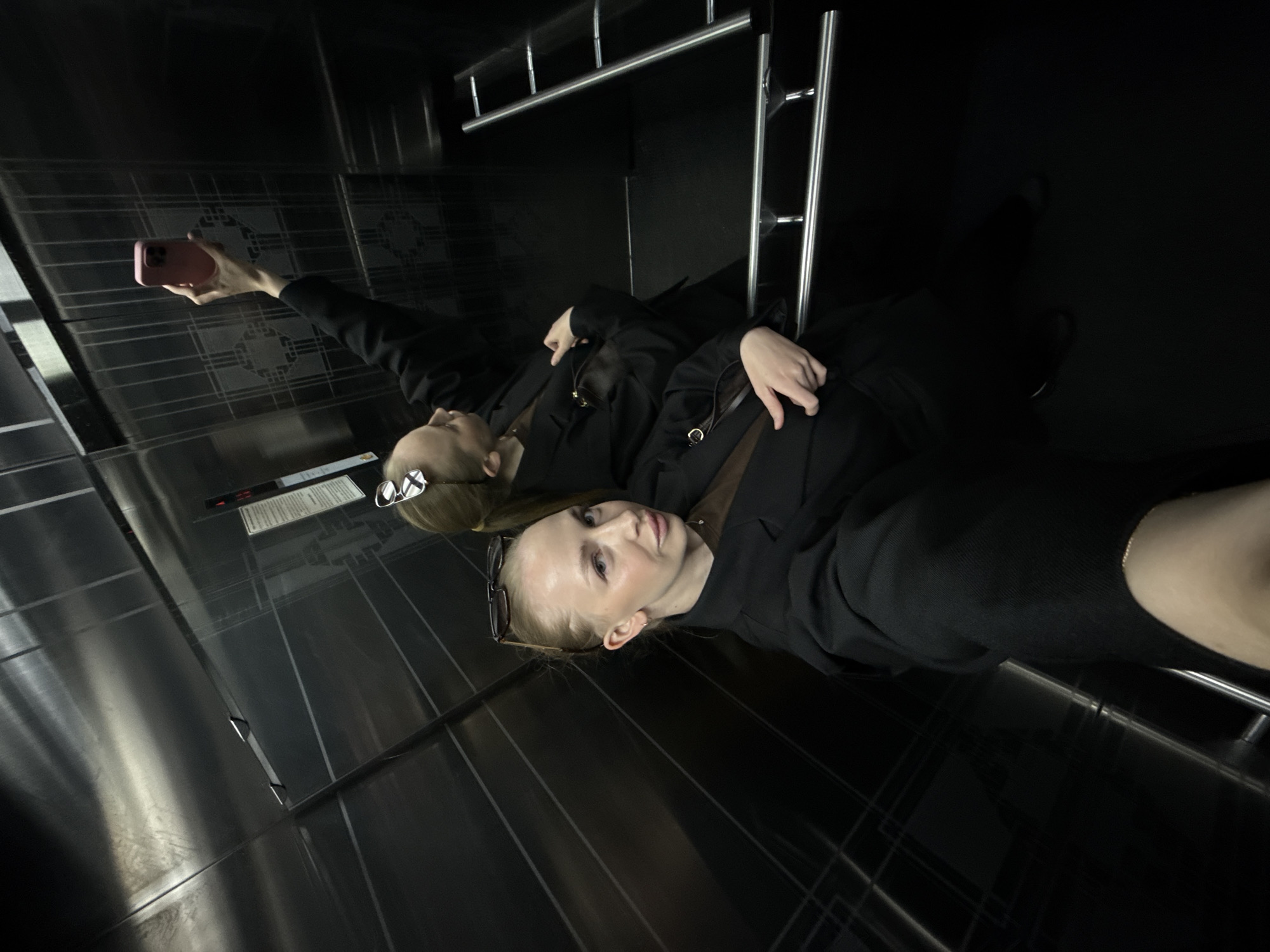 In an elevator with mirrored walls, a young woman with blonde hair pulled back and sunglasses perched on her head takes a selfie with her pink smartphone. She wears a dark blazer over a brown top and is reflected in the elevator's surface. Beside her, another woman, also with blonde hair, is seen from the waist up, holding her own pink smartphone to take a photograph of herself in the elevator's mirrored wall. She is wearing a similar dark blazer. The elevator's interior is dark, with silver railings and patterned metallic walls. The floor is black. The image is taken from a high angle. The location is Petropavlovsk-Kamchatsky, Russia.