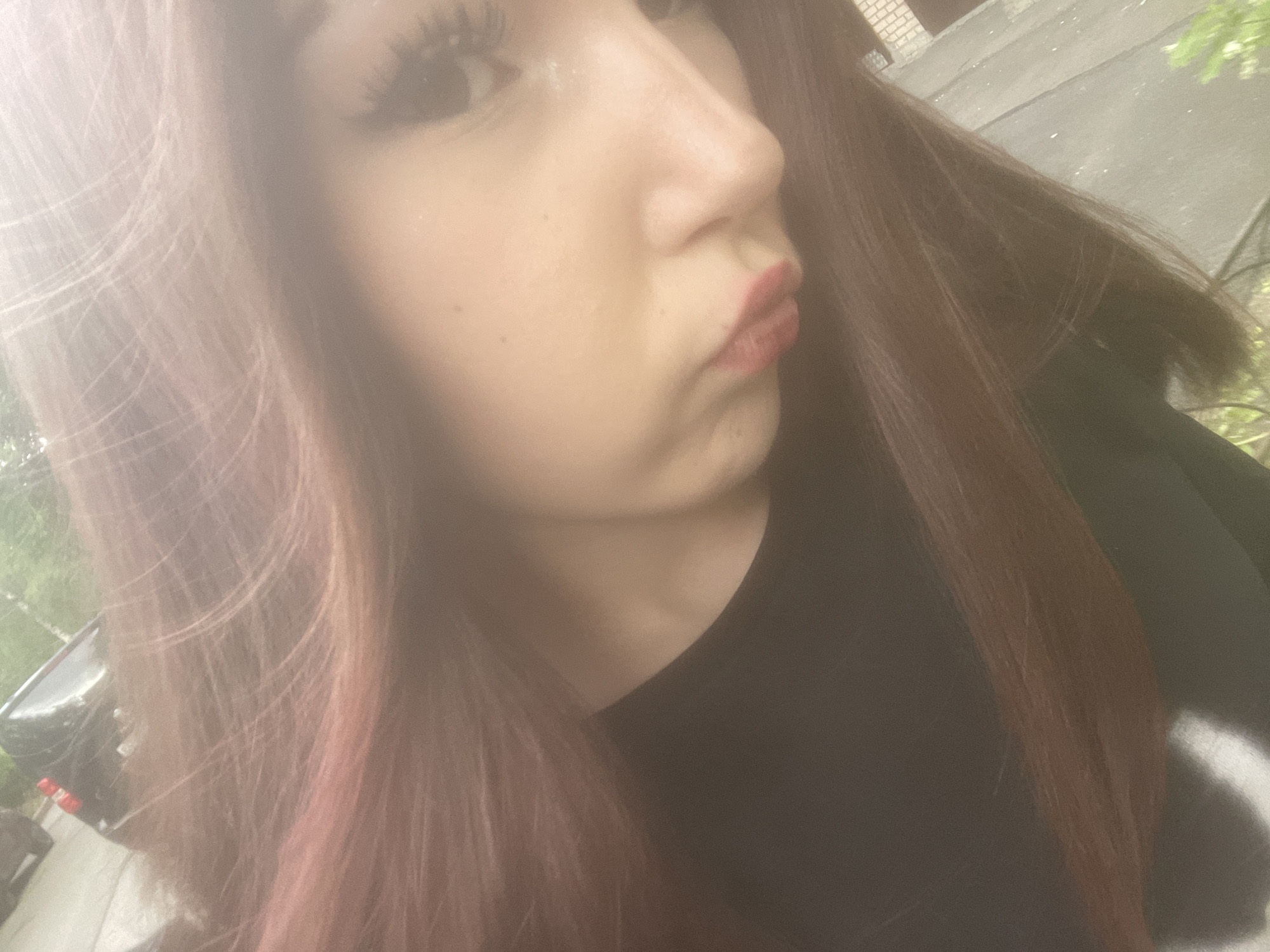 A close-up, slightly high-angle selfie captures the right side of a young individual's face, with her long, reddish-brown hair cascading around her. The person's eye, adorned with prominent eyelashes, looks slightly upward and to the left. Her nose is visible in profile, and her lips are pursed in a slight pout, possibly an intentional pose. She is wearing a dark, likely black, top.
The image has a soft, hazy, and diffused quality, giving it a dreamlike or artistic filter effect. This haziness, possibly due to light reflection or a lens effect, obscures some details and creates a luminous glow around the subject.
The background suggests an outdoor setting, though specific features are indistinct due to the shallow depth of field and haziness. Green foliage can be seen on the left and right, implying trees or bushes. A car's side mirror is partially visible on the far left. On the upper right, there appears to be a dark structure, possibly part of a building or a dark road surface with faint outlines that could be brickwork. The overall lighting indicates daytime. There is no visible text in the image. The general setting suggests an urban or suburban outdoor environment, consistent with a location such as Saint Petersburg, Russia.