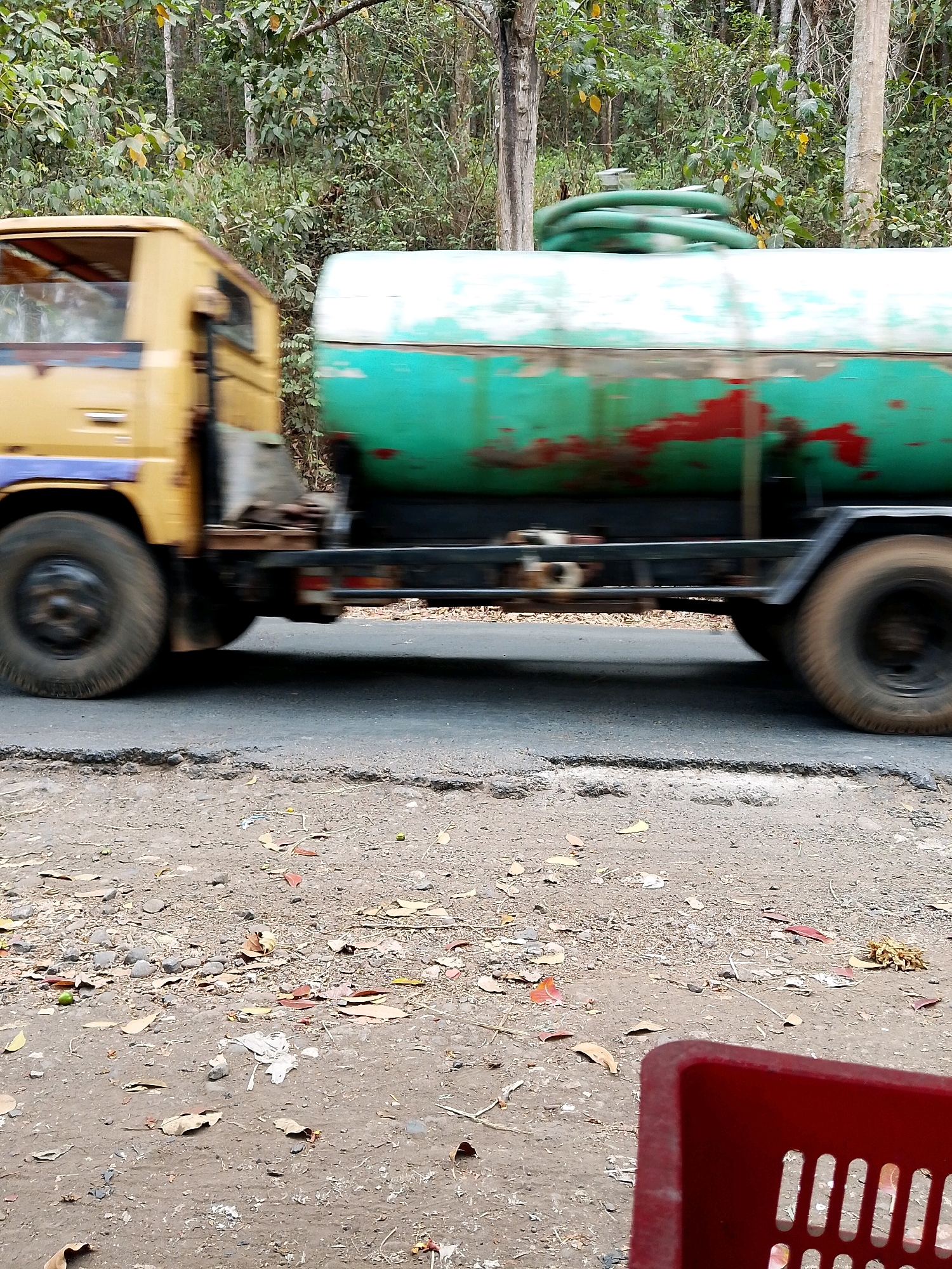 A yellow truck with a large green tank drives on a cracked asphalt road. The truck is in motion, indicated by motion blur on the wheels and the truck's body. The tank is predominantly green with patches of red showing through the paint. A coiled green hose is visible on top of the tank. The road is surrounded by a dense, leafy forest. The foreground shows a dirt shoulder with scattered leaves and pebbles. A red plastic basket is partially visible in the bottom right corner of the frame. The scene appears to be in a rural or natural area, possibly along a road in Pagak, Indonesia, given the vegetation and road conditions.