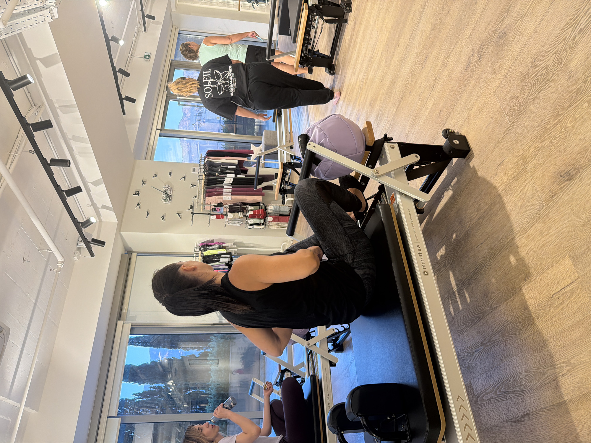 The image depicts an indoor Pilates studio with light wood flooring and large windows overlooking a natural landscape. The studio is equipped with multiple Pilates reformers. In the foreground, a woman with dark hair pulled back is seated on a reformer, wearing a black tank top and patterned leggings. She is holding her hands near her midsection. To her left, another woman with blonde hair, wearing a light purple top and dark leggings, is seated on a reformer and holding a bottle. In the background, two more women are visible. One, wearing a black t-shirt with "SOLEIL" printed on the back, is walking away from the camera. Another woman, wearing a light green tank top, walks past a rack of athletic clothing. The overall lighting is bright, with track lighting visible on the ceiling.