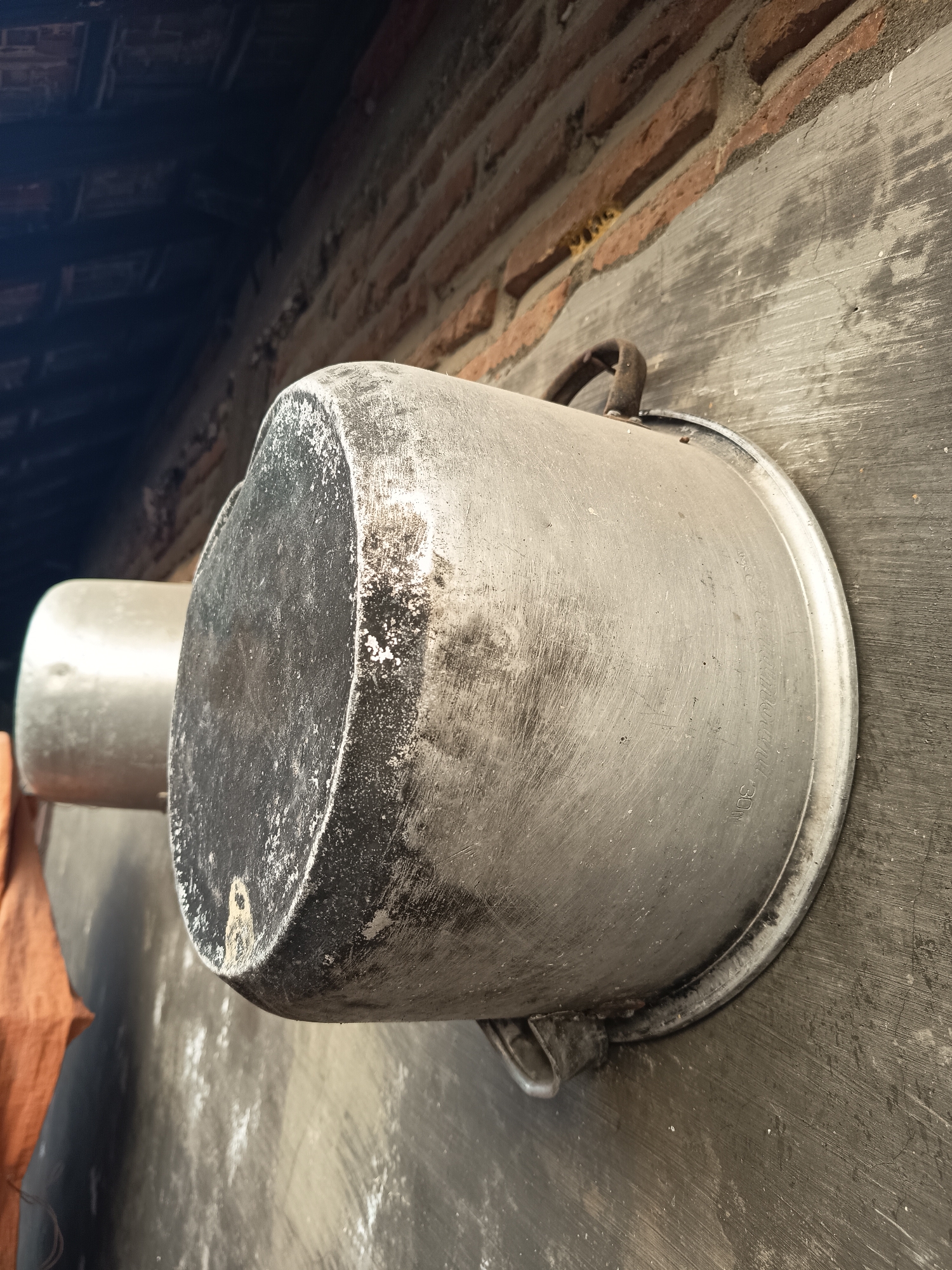 Two aged aluminum pots hang against a textured grey wall with a brick wall above. The larger pot, in the foreground, is positioned horizontally and appears to be a kettle or a large cooking pot, showing signs of wear and a dark, sooty residue around its opening. A rusty metal handle is attached to its side. Behind it, another similar pot is visible, appearing slightly lighter in color. The wall beneath the pots is rough and shows some vertical markings. Above the grey wall, a section of a brick wall is visible, constructed with reddish-brown bricks and grey mortar. The roofing structure, made of dark wooden beams, is visible in the upper left corner of the image, suggesting an outdoor or semi-enclosed area. The scene is likely in Sugio, Indonesia, given the context.