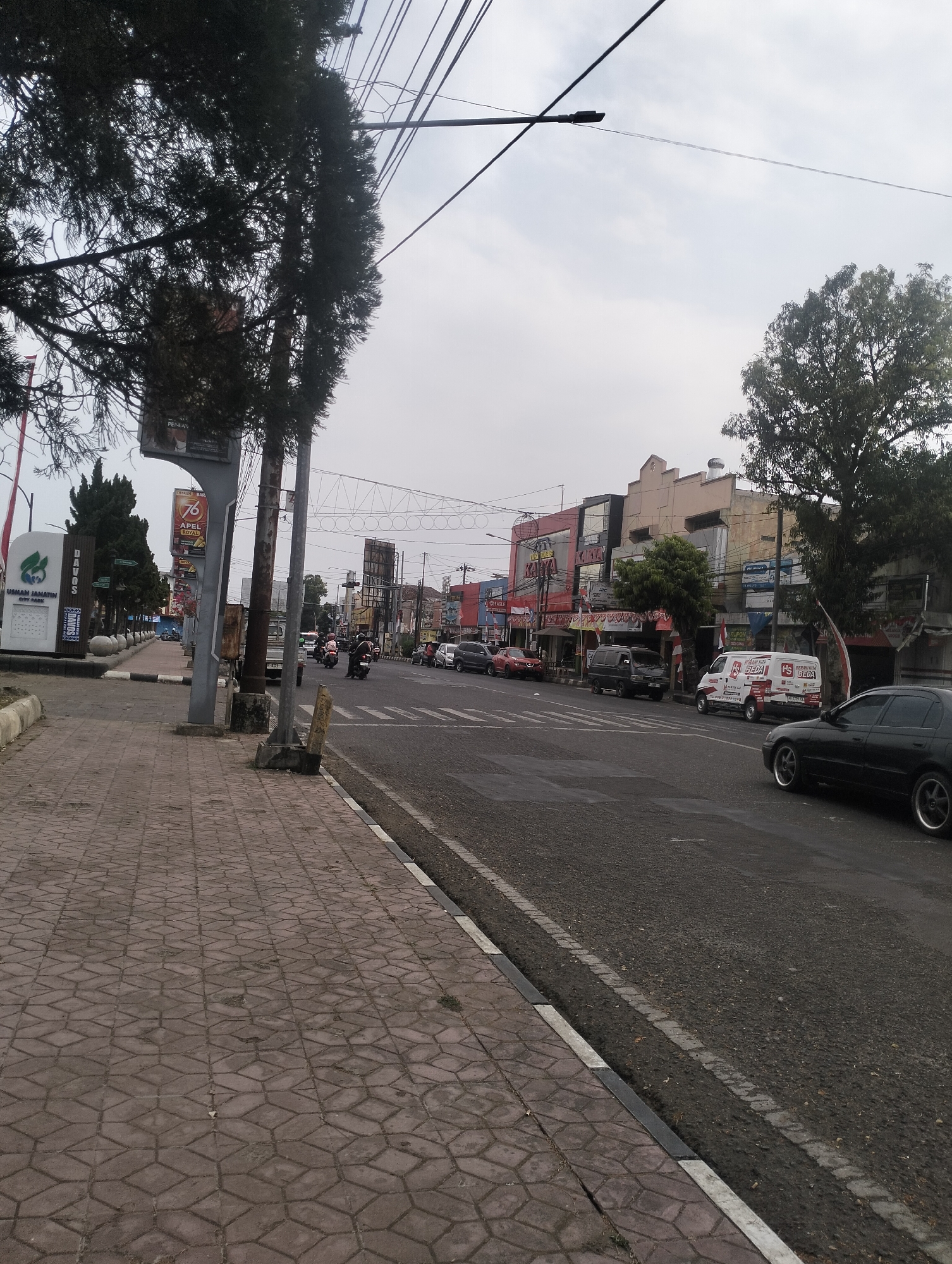 The image depicts a street scene in Purbalingga, Indonesia. The foreground shows a patterned sidewalk with a curb bordering a two-lane asphalt road. On the sidewalk, there are utility poles, trees, and signage, including a large sign that reads "DADOS USMAN JANIEN CITY PARK." Further down the sidewalk, there are bollards and more trees.
The road shows traffic, with several vehicles visible including motorcycles, cars, and vans. Some of the cars are parked along the side of the road. The buildings lining the street are a mix of commercial establishments, with various storefronts and signage. Some buildings have multiple stories and display Indonesian script. The sky is overcast. The overall impression is of an urban commercial area on a cloudy day.