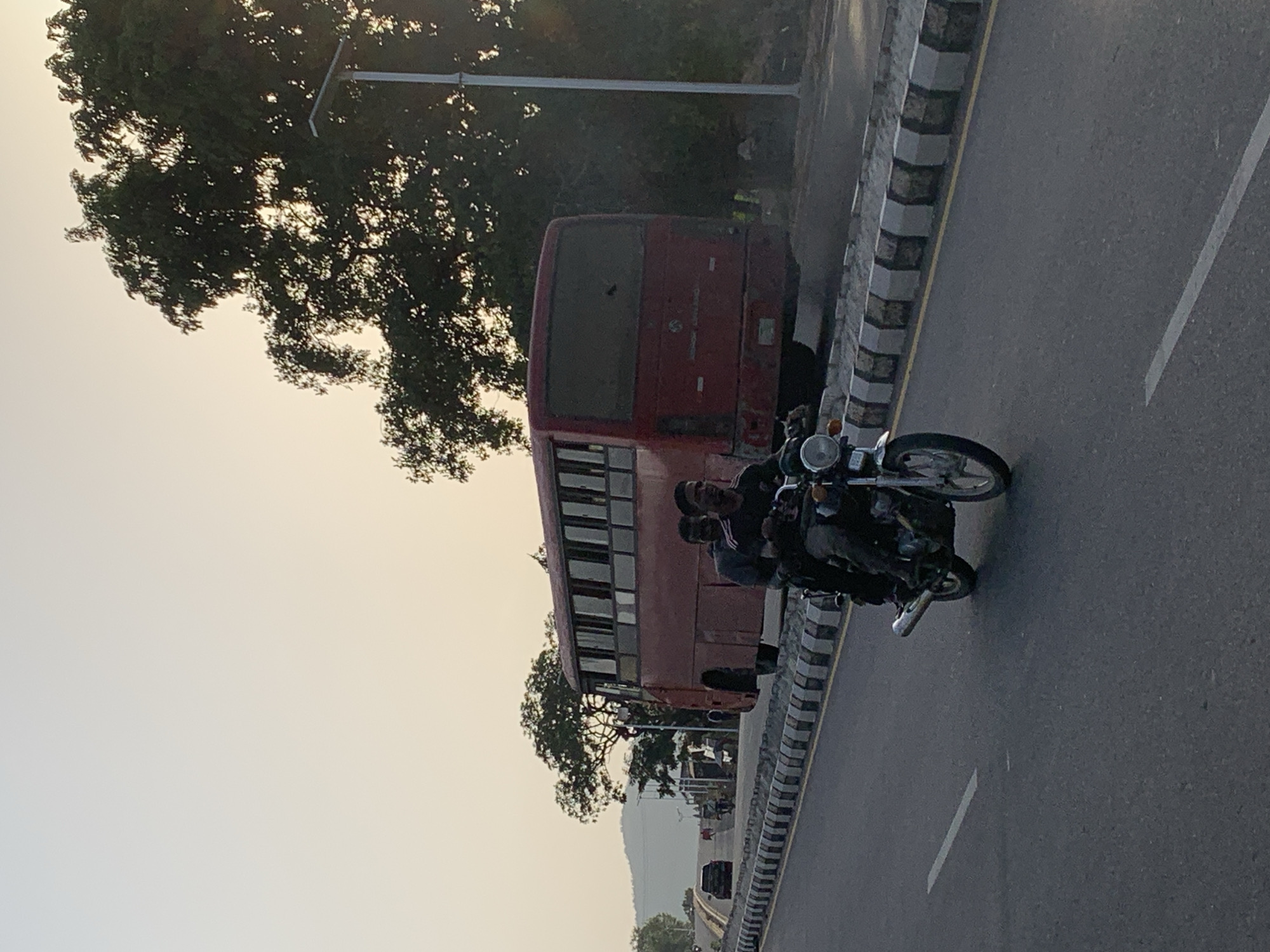 A red bus is parked on the side of a road with a motorcycle to its right and two individuals riding on the motorcycle. The road appears to be in Zango-ndaji, Nigeria. There are trees to the left of the bus and a mountain in the distance. The sky is bright, suggesting it is daytime.