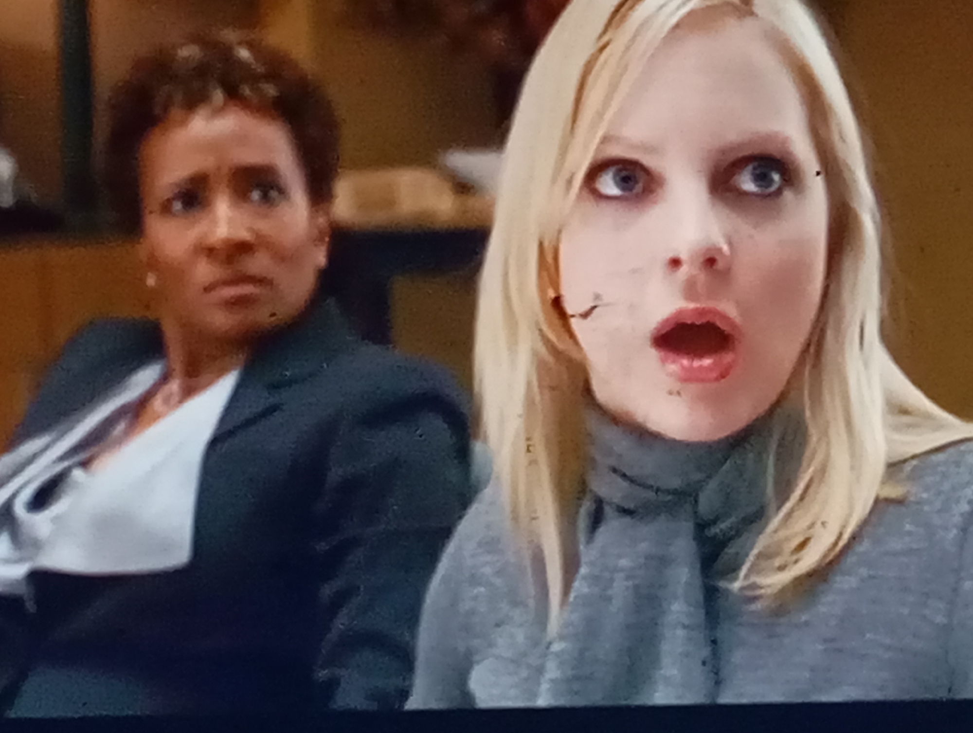 The image is a close-up, indoor shot showing two women. On the left, a Black woman with short, curly hair is looking to her right with a concerned expression. She is wearing a dark suit jacket over a white collared shirt. On the right, a white woman with blonde hair is looking forward with a surprised expression, her mouth open. She is wearing a grey turtleneck sweater. The background is blurred, suggesting an indoor setting with warm-toned walls and furniture. There are no visible indicators of the city of Sofiivska Borshchahivka, Ukraine, or any specific location.