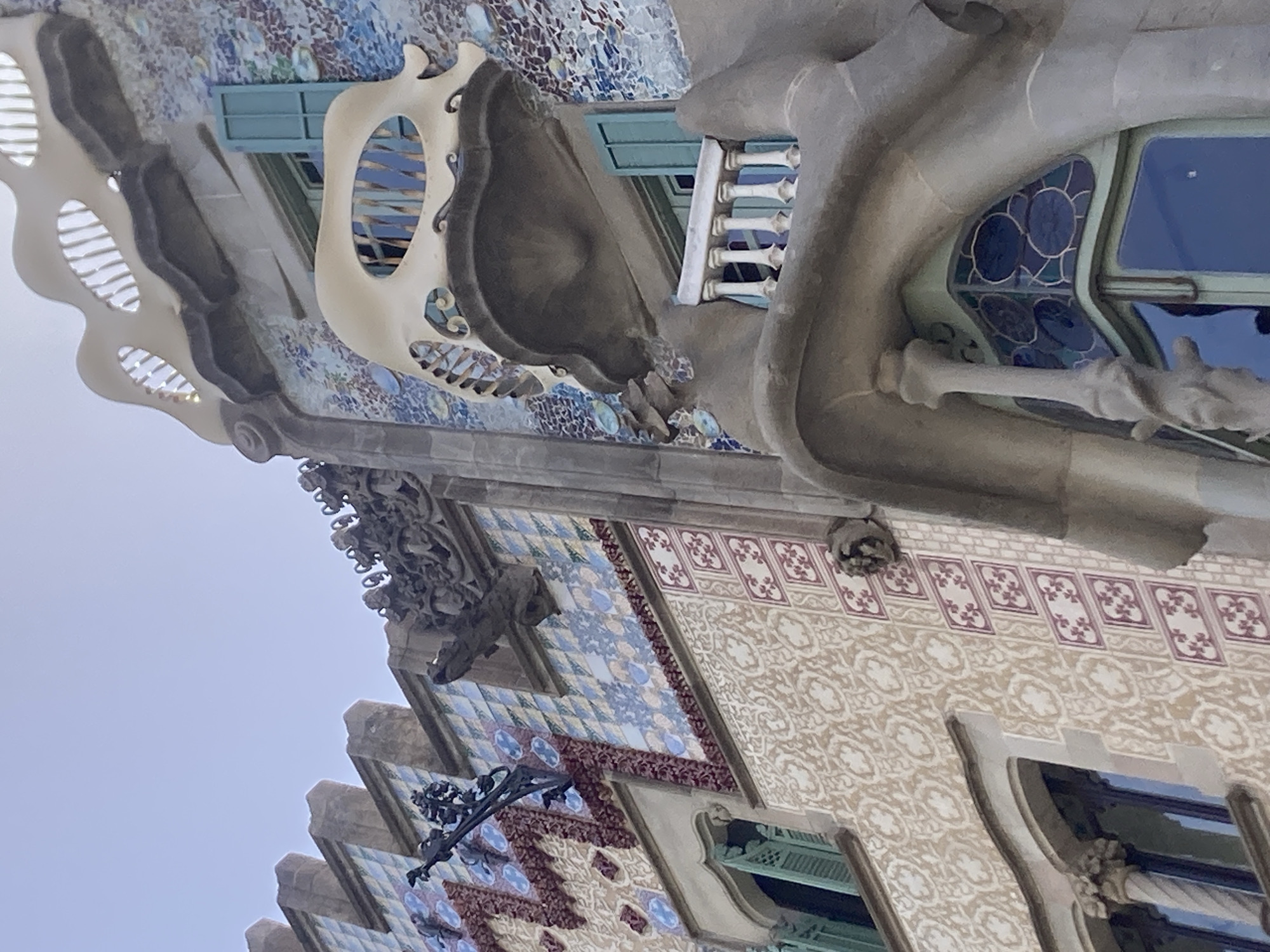 This is a photograph of the facade of Casa Batlló, a building located on Passeig de Gràcia in the Eixample district of Barcelona, Spain. The image focuses on the intricate architectural details of the building's exterior. The facade is adorned with a mosaic of colorful tiles, predominantly in shades of blue and white, with some red and brown accents. Several windows are visible, some with light blue shutters. The building features unique balconies, one of which has a bone-like structure and is shaped like a shield. Another balcony above it is more organic, with wavy lines and openings. Stone sculptures resembling gargoyles or mythical creatures are integrated into the upper sections of the building. The sky above is clear and light blue.