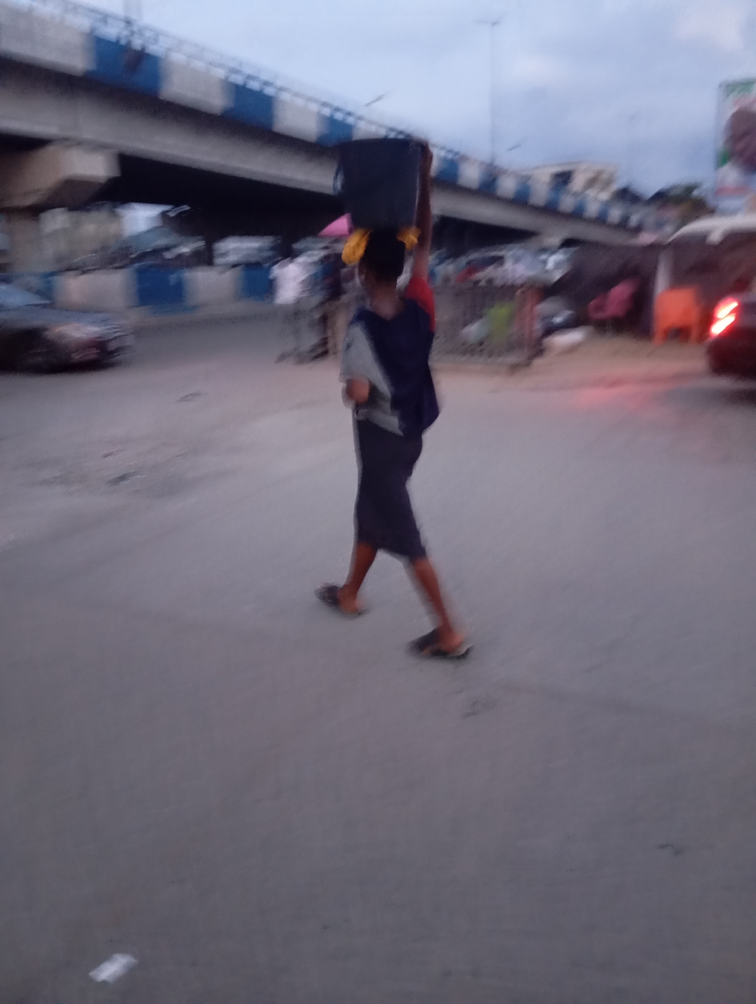 An individual, possibly female based on clothing and hair, walks carrying a large black rectangular object on their head. The person is wearing a navy blue top with red trim and a dark skirt. They are also wearing flip-flops. The background features a concrete overpass with blue and white stripes, suggesting an urban environment. In the distance, there are some blurred structures and vehicles, including a red tail light from a vehicle to the right. The ground appears to be paved asphalt. The scene is dimly lit, indicating it might be dusk or dawn. The overall impression is that of street life in an African city, potentially Port-Harcourt, Nigeria, given the visual cues of the overpass and the style of dress and activity.
