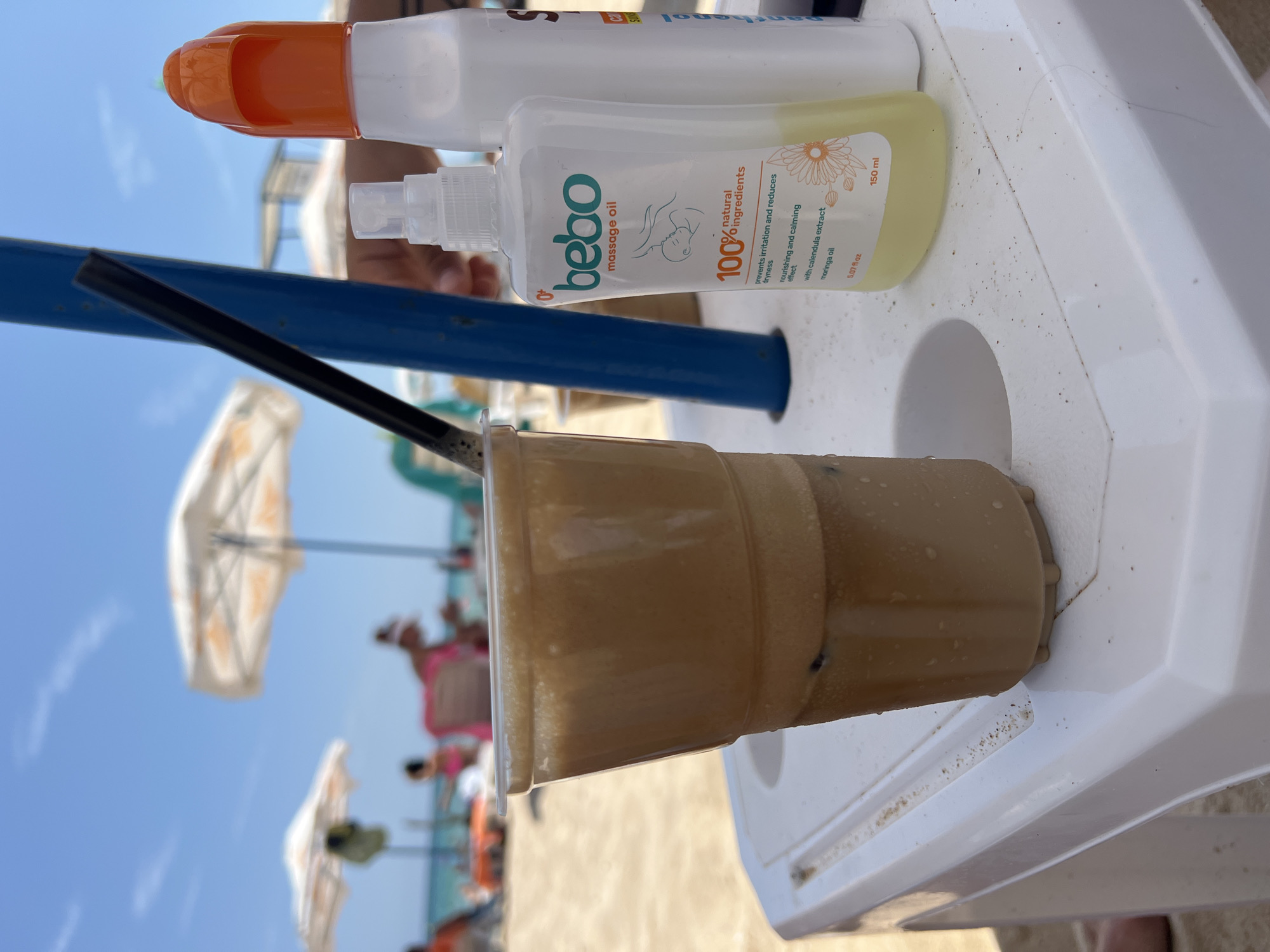The image shows a close-up view of a plastic cup filled with a foamy, brown beverage, likely a coffee drink like freddo cappuccino, with ice cubes visible. A black straw extends from the cup. The cup rests on a white plastic table. To the right of the cup are two bottles: a white spray bottle labeled "bebo massage oil" with orange text and a smaller white bottle with an orange cap. In the background, a beach scene unfolds with blurred figures of people relaxing under beach umbrellas. The environment appears to be a sunny beach, possibly in Primorsko, Bulgaria, given the style of the umbrellas and the general setting. The focus is primarily on the beverage and the products on the table, with the beach serving as a backdrop.