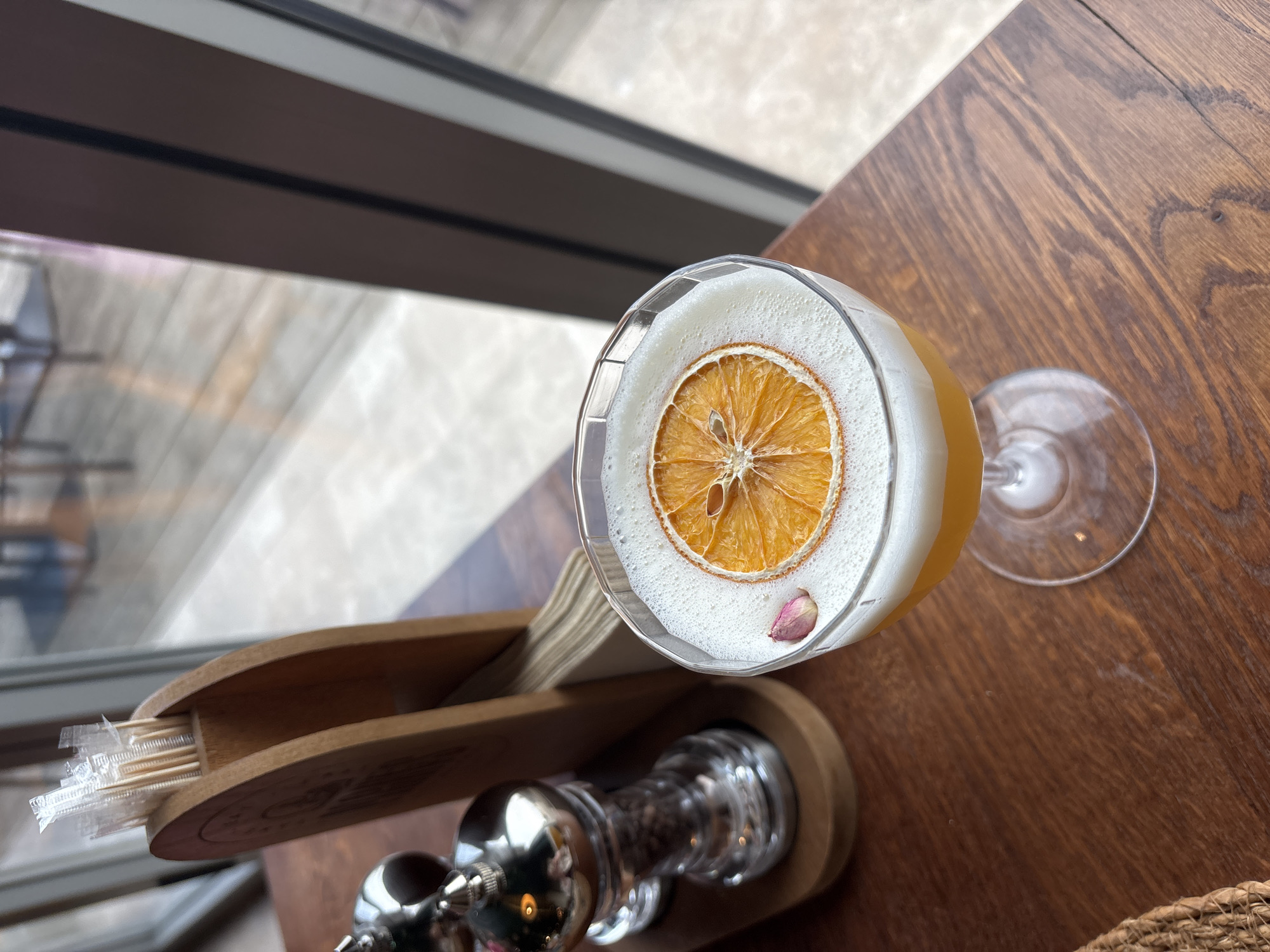 The image shows a cocktail on a wooden table, next to a toothpick holder and a window. The cocktail is garnished with a dried orange slice and a small rose petal. The toothpick holder is made of wood and has a silver mechanism on top. The window looks out onto a city street. The location is likely in Varna, Bulgaria.