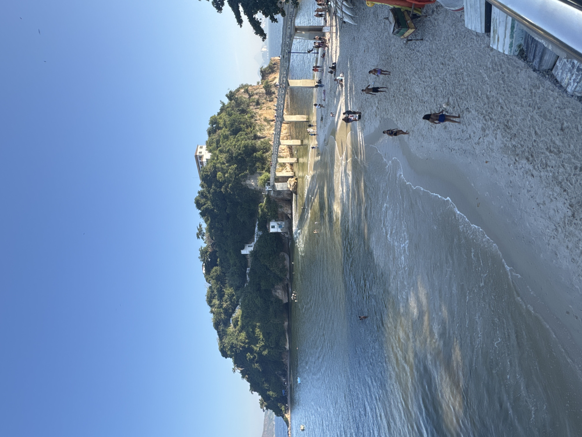 This is a photograph of Icaraí Beach in Niterói, Brazil, taken on a sunny day with a clear blue sky. The beach is populated with people engaging in various activities. Some individuals are sunbathing on the sand, while others are walking along the shoreline. Several people are visible in the ocean, swimming or wading in the water. A prominent feature in the image is a long pedestrian bridge extending from the sandy beach towards a rocky, tree-covered island. There are buildings visible on top of the island and along the shoreline. In the foreground, there are some boats and other beach equipment. The overall scene depicts a lively and picturesque beach environment.