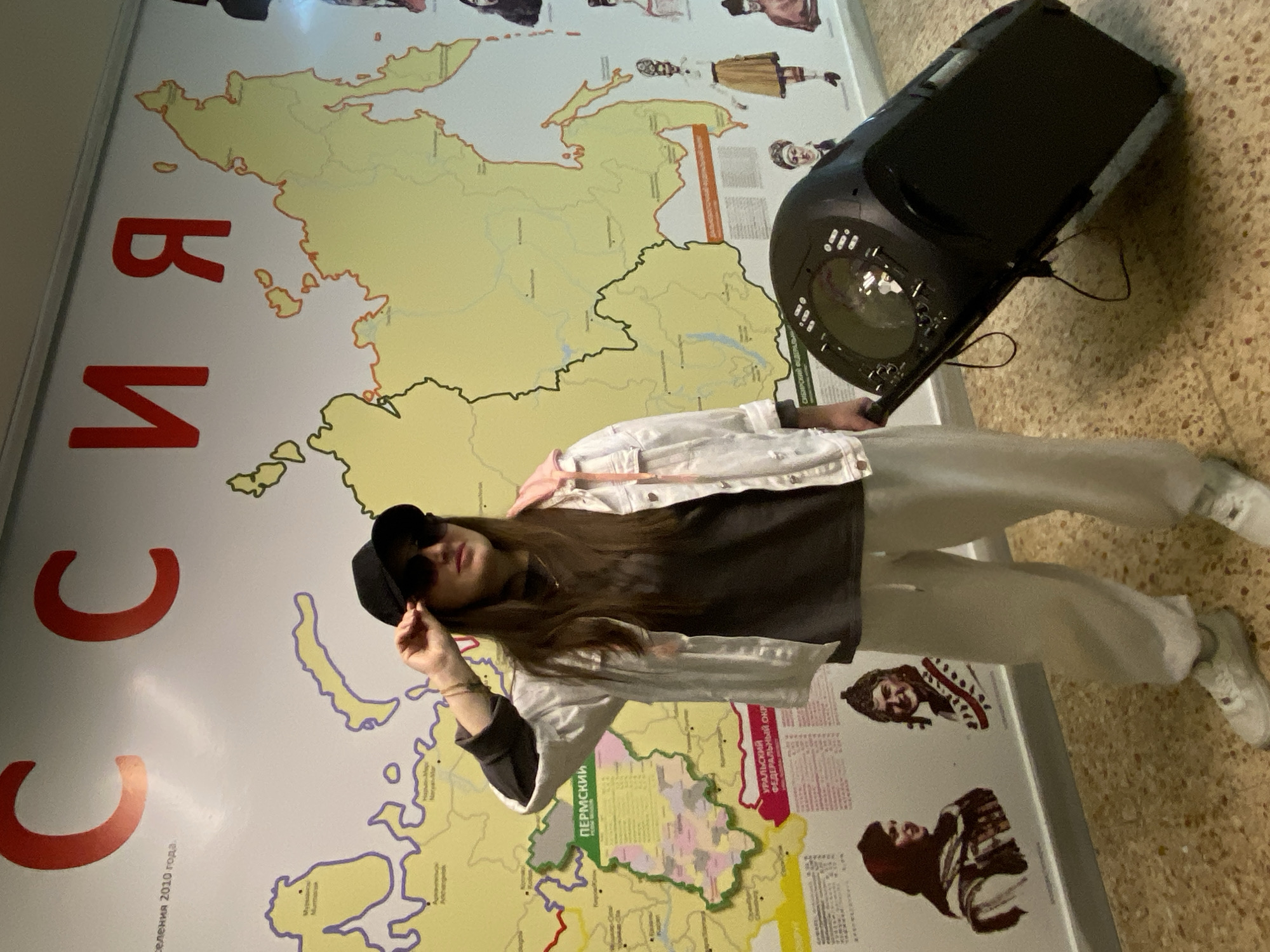 An indoor scene shows a young person standing next to a large wall map of Russia. The individual, who appears female, has long dark hair and is wearing a black baseball cap, sunglasses, a white denim-style jacket over a dark shirt, light-colored pants, and white sneakers. In her right hand, she holds a large black portable speaker with a visible, illuminated front panel.
The background is dominated by a detailed, light-yellow map of Russia, outlined with red and black borders. Prominent red Cyrillic letters on the left side of the map spell "РОССИЯ" (RUSSIA). Other legible Cyrillic text includes "2010 года" (2010 year), "ПЕРМСКИЙ" (Permsky), and "УРАЛЬСКИЙ ФЕДЕРАЛЬНЫЙ ОКРУГ" (Ural Federal District). The map also features small illustrations depicting people in various traditional attire. The floor is a light-colored, speckled tile. The person is posing casually, with one hand touching the brim of her cap, exuding a confident and relaxed demeanor. The presence of the Russian map and Cyrillic text strongly suggests the location is in Russia, specifically Markovskii as provided.