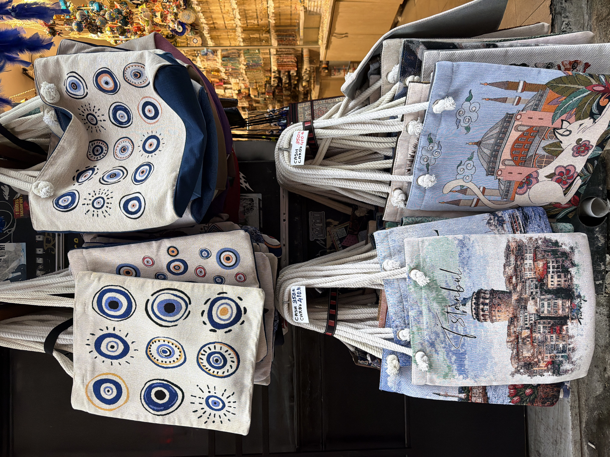 The image shows a display of tote bags hanging outside a shop. The bags are made of a woven fabric and feature various designs. Some bags have a pattern of evil eye symbols in blue and white. Others have printed designs of Istanbul landmarks, such as the Galata Tower and Hagia Sophia, one of which has a cat motif. A price tag is visible on one of the bags, indicating "CASH 350tl" and "CARDS 410tl". The background suggests an outdoor market or street scene, with shelves filled with other goods, possibly souvenirs, visible in the distance. The lighting is natural, suggesting daytime. The location is identified as Kadıköy, Turkey.