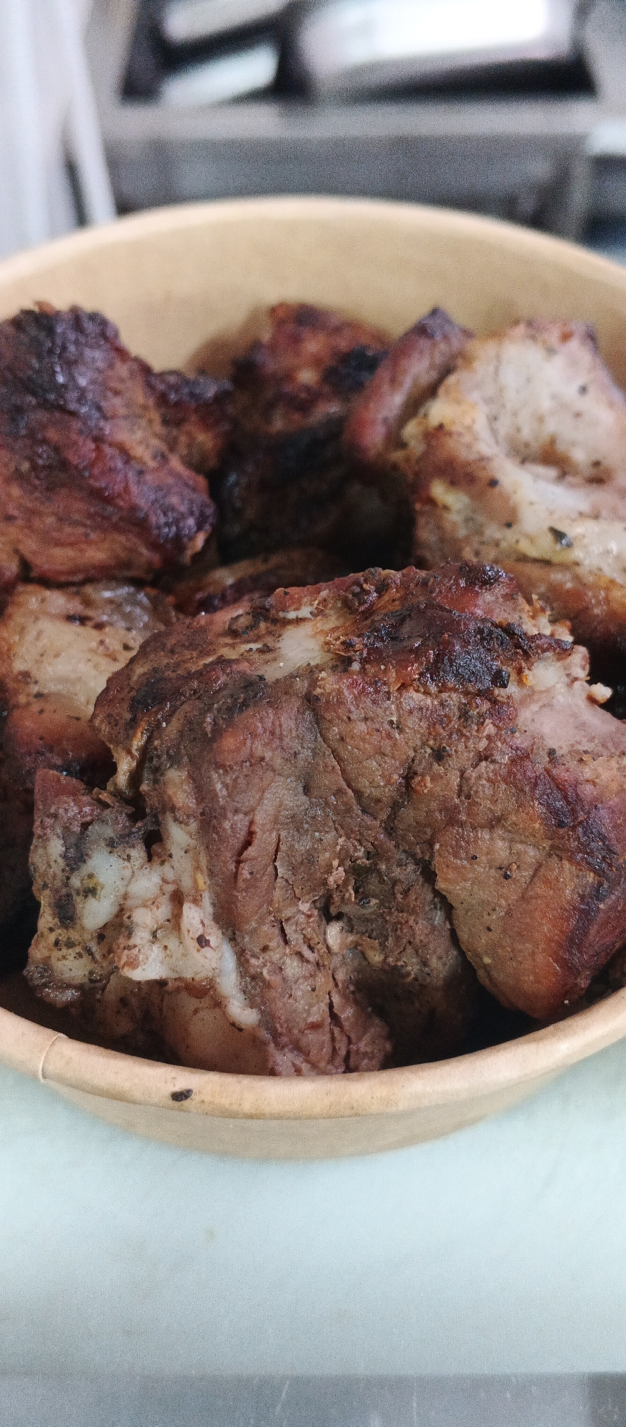 In Ploske, Ukraine, a close-up, eye-level shot captures a bowl filled with cooked meat. The meat appears to be pork, with visible layers of fat and muscle tissue, and a dark, seared exterior indicative of grilling or roasting. The bowl itself is a light brown, disposable container, likely made of paper or cardboard, with a slightly worn edge. The background is blurred, showing a metallic surface and an out-of-focus kitchen environment. The lighting is natural and comes from above, highlighting the texture of the meat.