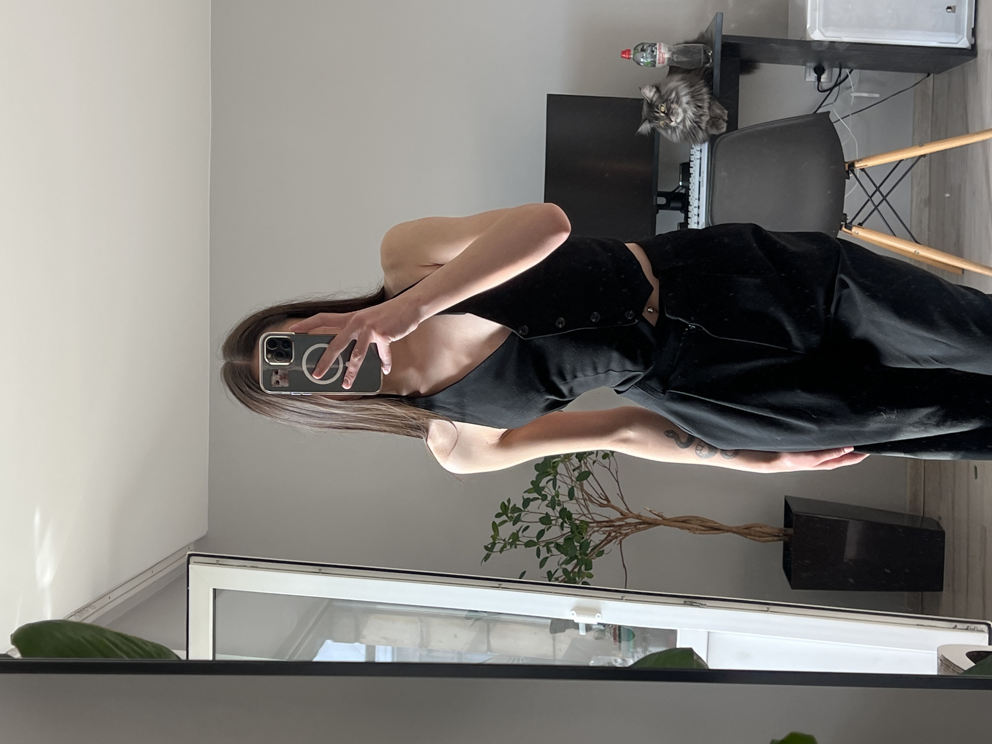 A person is taking a mirror selfie in a room. The individual is wearing black clothing, including a top with buttons and pants. A smartphone is visible in their hand, used to capture the image. The room contains a window with visible outdoor scenery, a potted plant, a desk with a monitor and keyboard, a chair, and a cat sitting on the desk near a water bottle. The wall is light gray. The image provides no explicit information confirming it was taken in Ternopil, Ukraine.