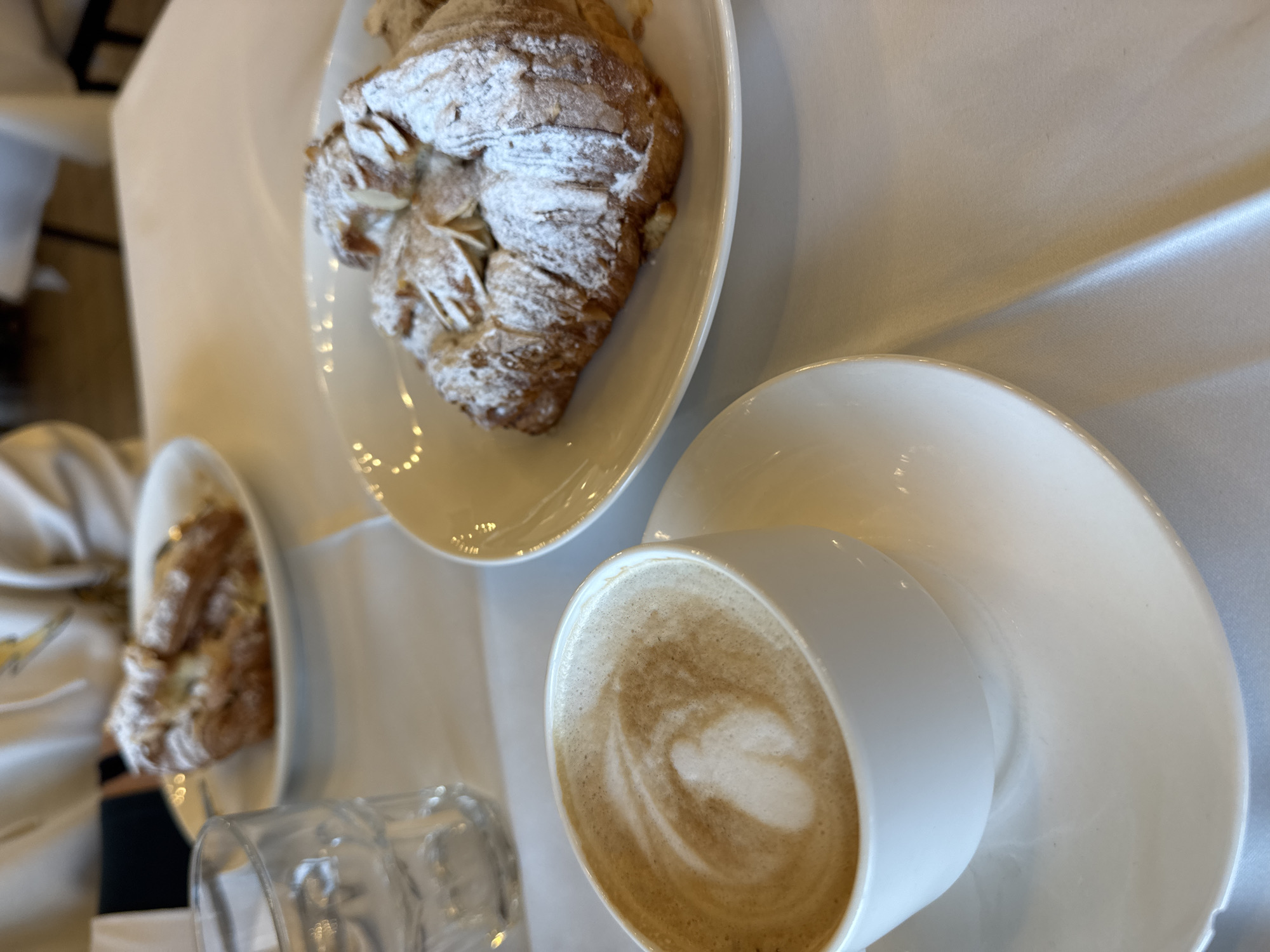 The image shows a table setting in an Unknown, Unknown cafe. There is a white tablecloth, and a plate of a sugar-powdered pastry and another plate of a plain pastry are visible. There is a cup of coffee sitting on a saucer, and a glass next to the cup. The image is taken at an oblique angle, suggesting it was taken at eye level by someone sitting at the table.