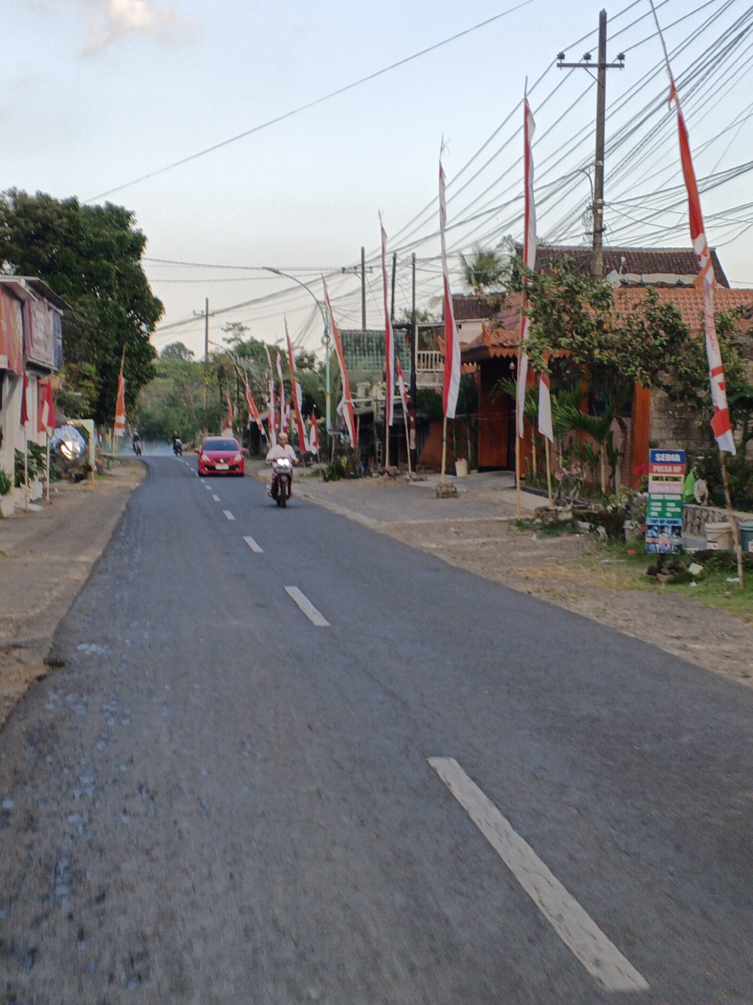 The image shows a road in Songgon, Indonesia, lined with Indonesian flags. The road is asphalt and has a white dashed line in the center. On the left side of the road, there are buildings and trees. On the right side of the road, there is a house with a tiled roof and more trees. There are several people on motorcycles on the road. One person is riding a motorcycle in the foreground, and another person is riding a motorcycle in the distance. A red car is also visible on the road. The sky is clear and blue.