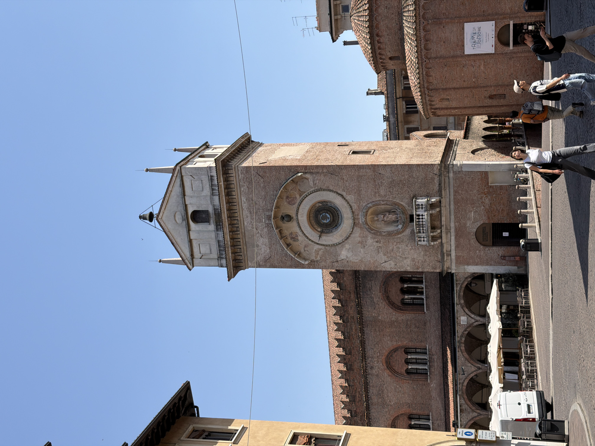 The image shows the Torre dell'Orologio in Mantua, Italy, set against a clear blue sky. The tower, constructed of brick with an ornate astronomical clock, dominates the scene. A group of four people are walking nearby. A cafe with outdoor seating is located at the base of an adjacent building, with a white van parked nearby. The scene is oriented vertically.