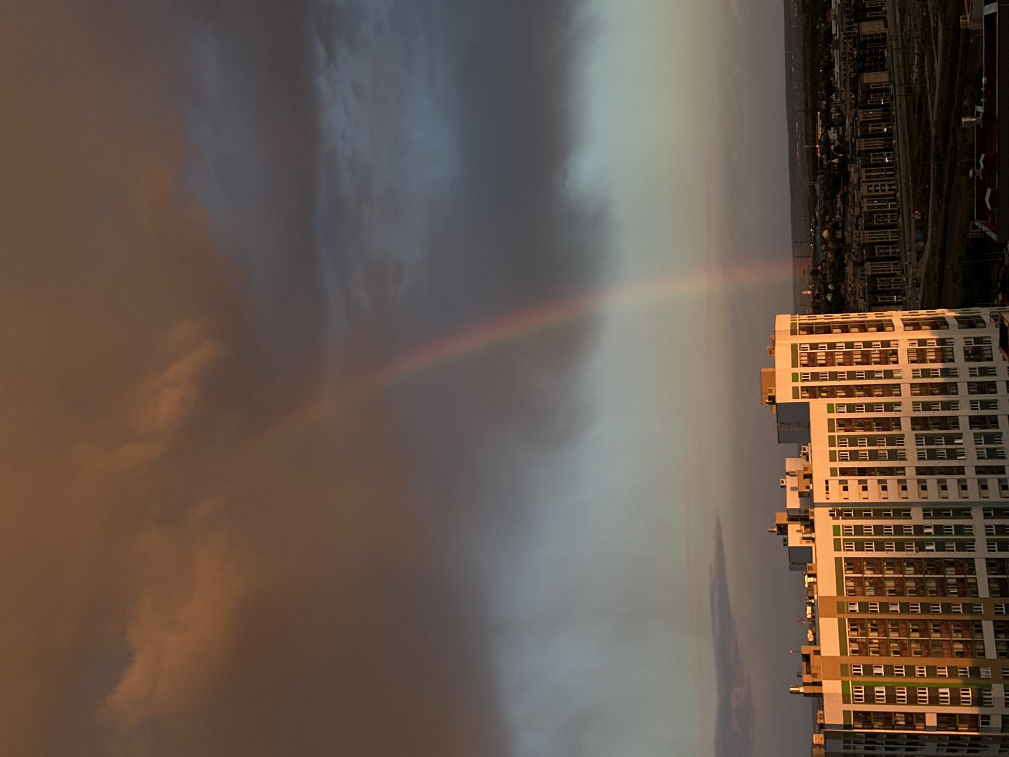 The image shows a rainbow over Yekaterinburg, Russia. A tall, multi-story building is located in the foreground on the right side. In the distance, a cityscape stretches along the horizon. Dark, ominous clouds dominate the sky on the left, while the right side of the sky is lighter and more overcast. A faint rainbow arcs across the middle of the image, connecting the building to the clouds.