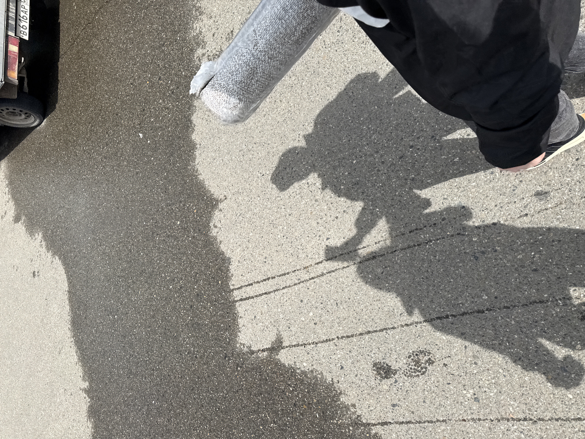 The image captures a section of paved asphalt, partially wet, with a vehicle visible on the left edge bearing the license plate "B616AP." Next to the vehicle lies a rolled, gray item, possibly a rug or carpet, wrapped in clear plastic. A person's lower torso, clad in black pants and a dark jacket, is also visible. The person's shadow, cast on the pavement, indicates they are holding the rolled item. The overall scene appears to be set in an urban environment.