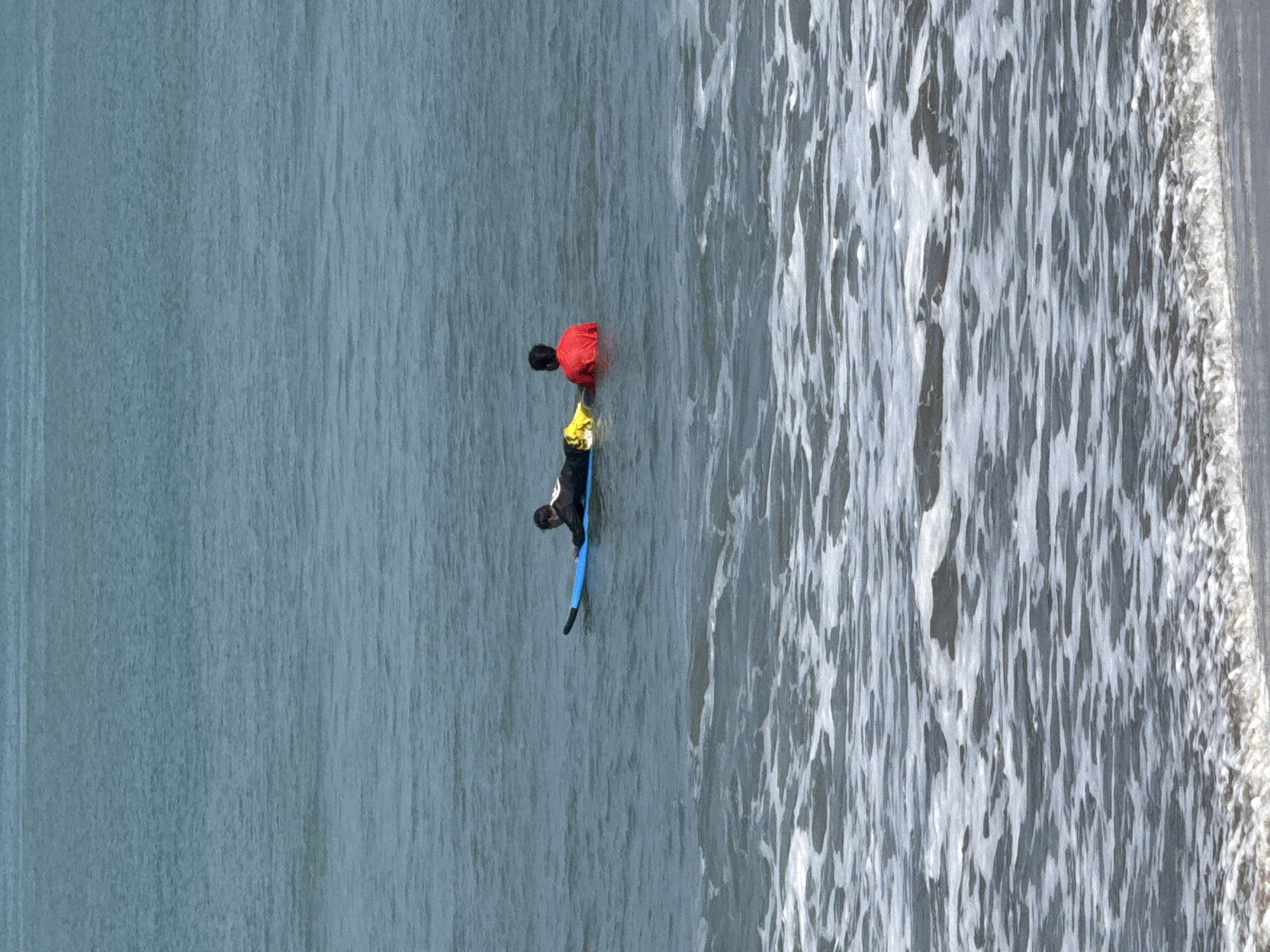 Two individuals are in the ocean in Kuta, Indonesia, on a blue surfboard. One person, wearing a black wetsuit, is lying prone on the surfboard, facing away from the camera. The second person, wearing a red shirt, is sitting on the surfboard behind the first person, also facing away from the camera. The surfboard is partially submerged in the water. White foamy waves are breaking closer to the shore, and the ocean is a muted blue-green. The scene appears to be a calm day at the beach.