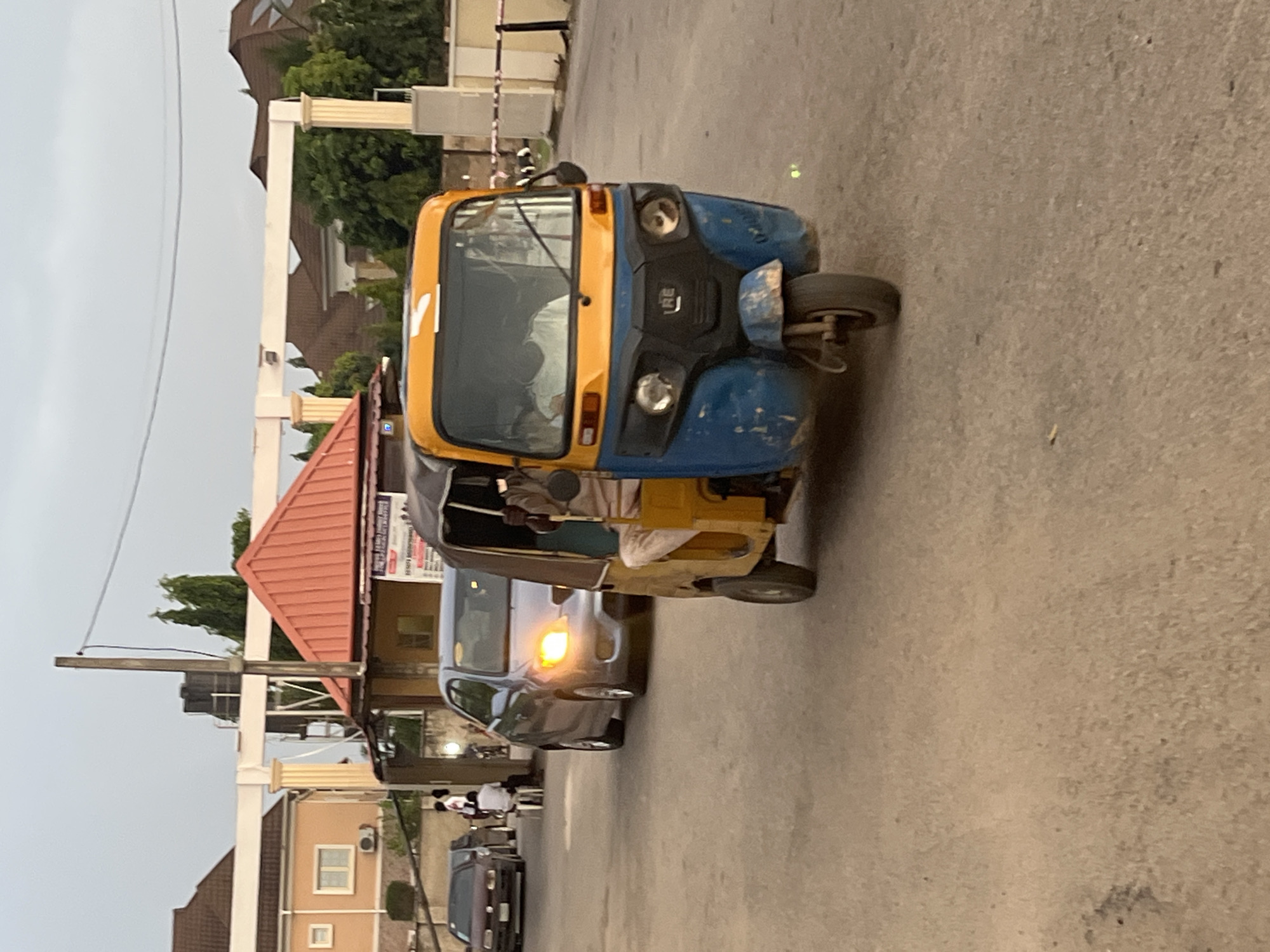 The image shows a blue and yellow motorized tricycle, commonly known as a "Keke Napep," parked on a paved street. A silver car is partially visible behind it. In the background, there are buildings, including one with a red roof and signage. Several people are visible on the sidewalk near the buildings. Based on the presence of Keke Napeps and the general architecture, the location is likely in a residential or commercial area in the city of Abuja, Nigeria.