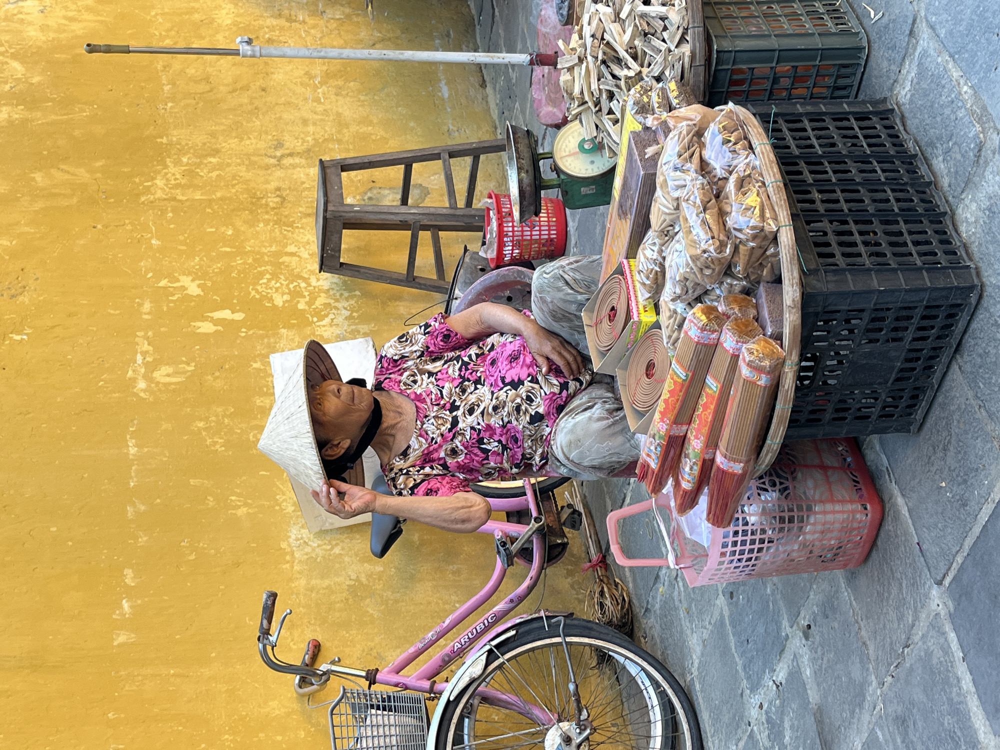 An elderly woman with light skin wearing a conical Asian hat and a patterned floral shirt is sitting on a pink bicycle in Hội An Ward, Vietnam. The woman appears to be resting. The bicycle has a basket on the front handlebars and a larger round basket filled with incense and other items attached to the back. The bicycle is parked next to a textured yellow wall on one side and a stone-paved area with black plastic crates on the other. A set of wooden steps, a green scale and more crates are nearby. The woman's hat and bicycle suggest she is a street vendor.