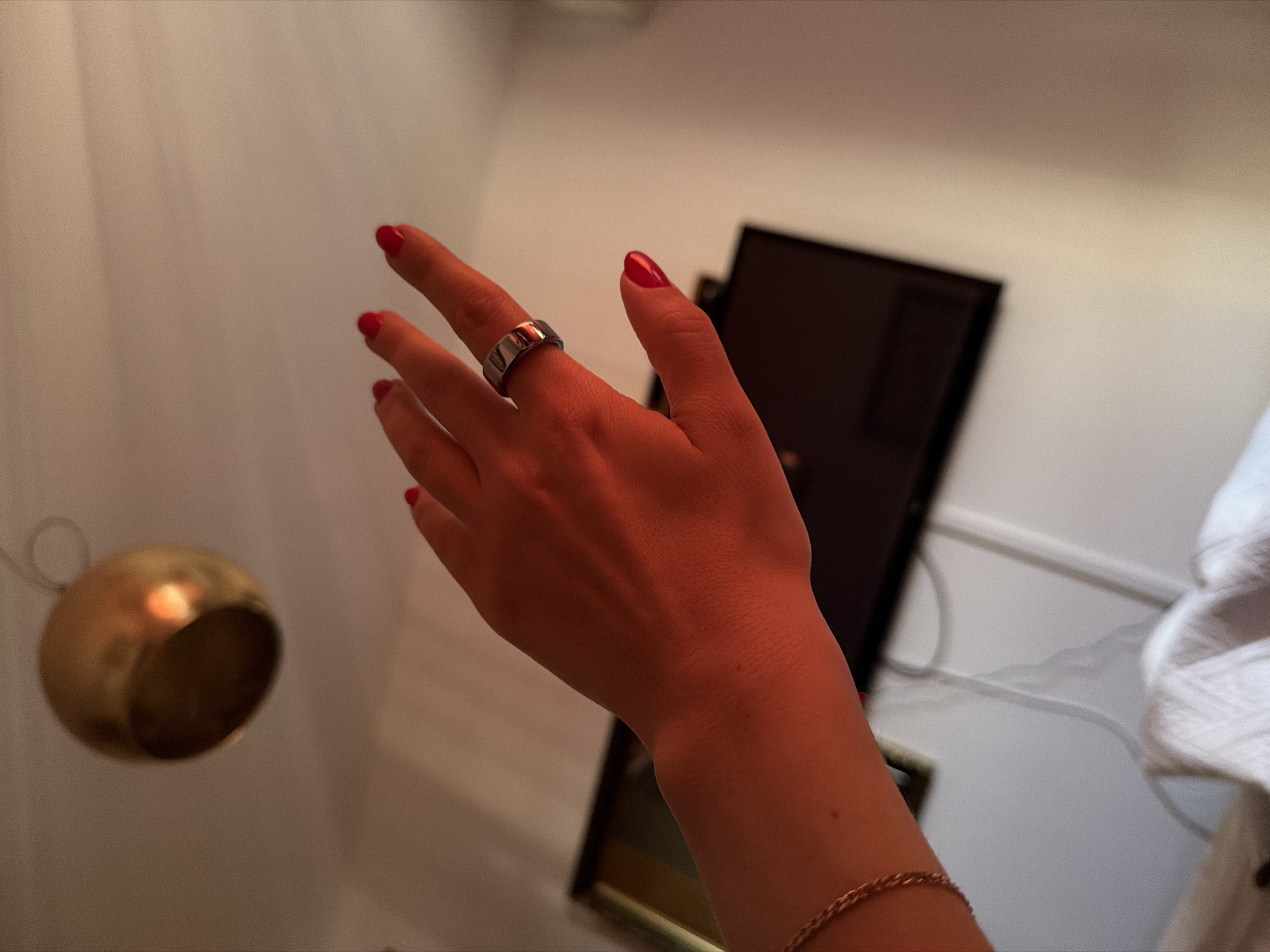 An indoor, close-up photograph captures a person's outstretched right hand, illuminated by warm, reddish light. The hand is positioned centrally, slightly angled, with fingers extended. The visible fingernails are painted with bright red nail polish. A wide, metallic, silver-toned ring adorns the ring finger. A thin, gold-colored chain bracelet is visible on the wrist.
The background primarily consists of a light-colored, possibly white or off-white, wall, which appears slightly textured. To the left of the hand, a blurry, golden, spherical object is visible, appearing to hang from a wire or loop. To the right and behind the hand, a dark, rectangular, reflective surface is partially visible, which could be a mirror, a framed piece of art, or an electronic screen. Further right, a white cable and a hint of white fabric (possibly a towel or clothing) can be seen against the wall.
The warm, dim lighting suggests an evening or night setting, or an indoor space with specific ambient lighting. No specific activity is clearly depicted beyond the extended hand, which might be posing or reaching for something out of frame. There is no visible text in the image. The location context for this media file is Budva, Montenegro.