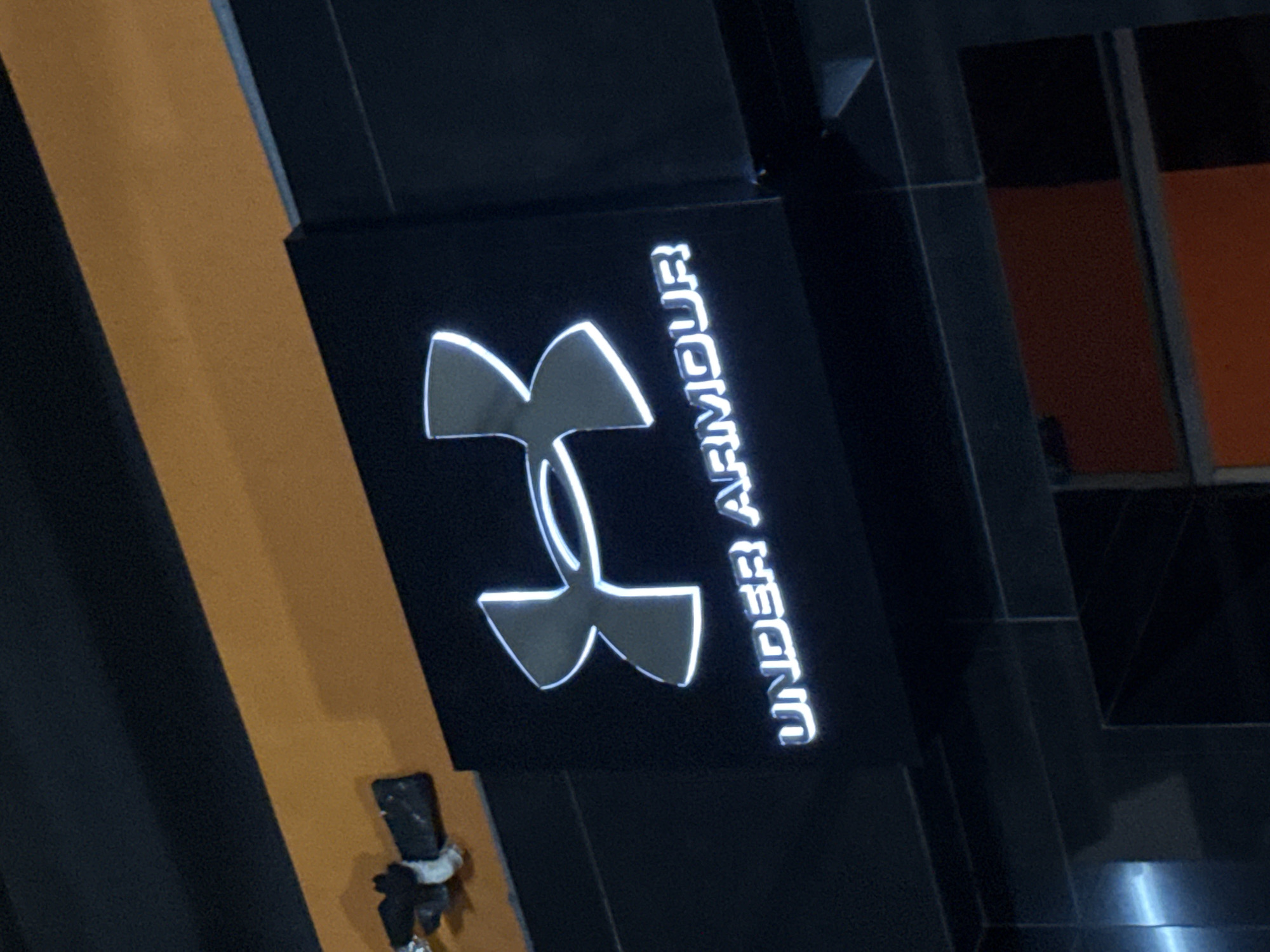 The image shows a close-up of a black sign with the "Under Armour" logo illuminated in white. The logo consists of a stylized "U" shape and the brand name written vertically next to it. The sign is mounted on a dark, angular surface, possibly part of a building or an interior design element. To the left of the sign, there is a diagonal stripe of bright orange against a dark background, creating a strong visual contrast. A small, dark object with white ties, resembling a piece of attached equipment or decoration, is visible hanging near the bottom left of the sign. The background features dark, textured surfaces and a glimpse of an orange-lit area, possibly a window or another illuminated section of the environment. The overall lighting suggests it might be indoors or at night, with the illuminated logo being the primary light source in the immediate area. The scene appears to be a commercial setting, likely a store or an athletic facility, given the brand displayed. There are no people visible in the image. The text "UNDER ARMOUR" is clearly legible. The image provides a detailed view of the brand's signage and its immediate surroundings.